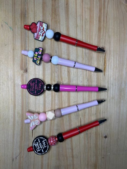 Five decorated pens with beads and charms arranged on a wooden surface.