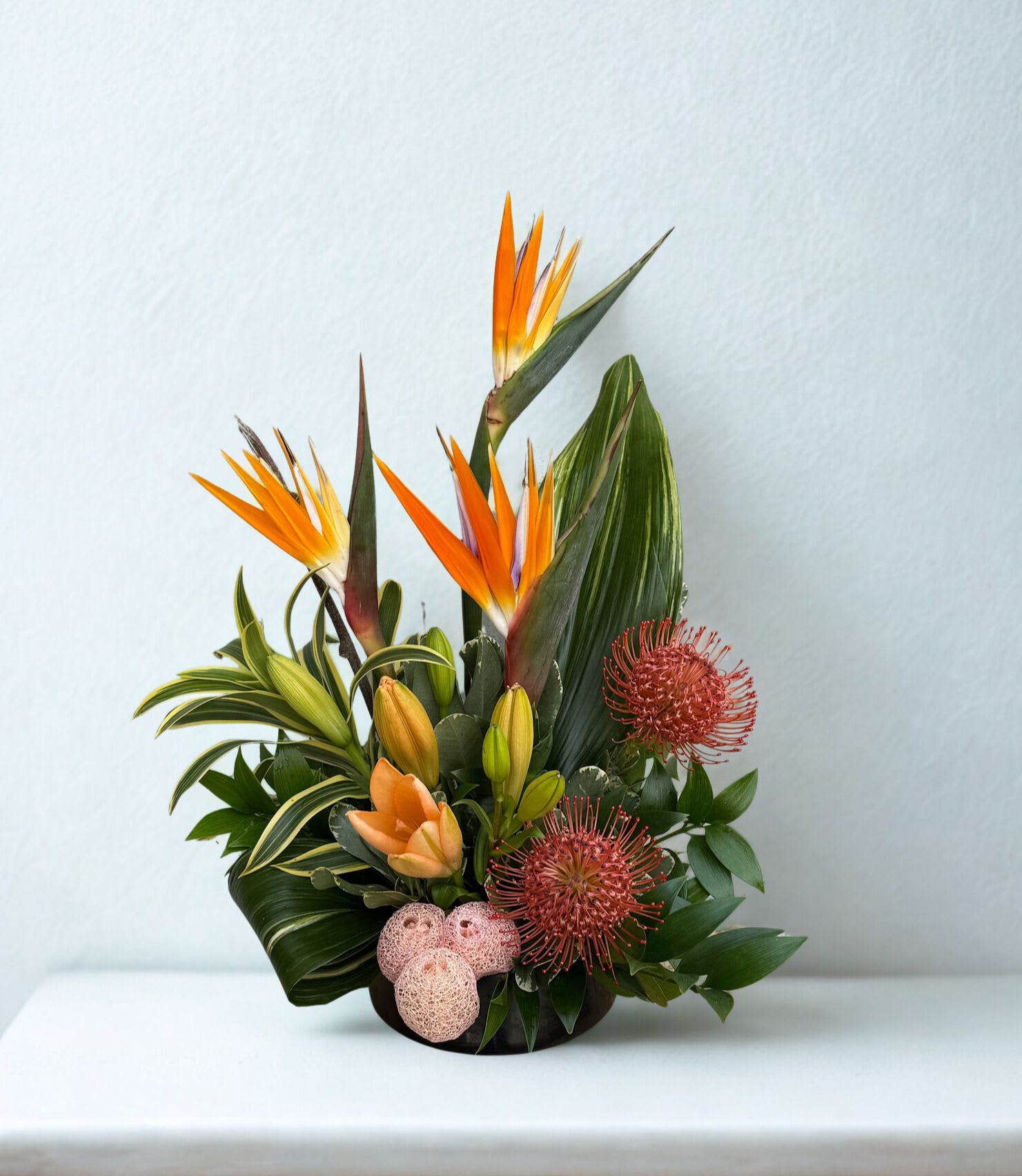 Gleaming Getaway - Bold, modern, and undeniably striking, this tropical arrangement brings a touch of the exotic indoors. Featuring vibrant birds of paradise, textured pincushion protea, and warm-toned lilies, it’s artfully designed with lush greenery for a sculptural, contemporary look.