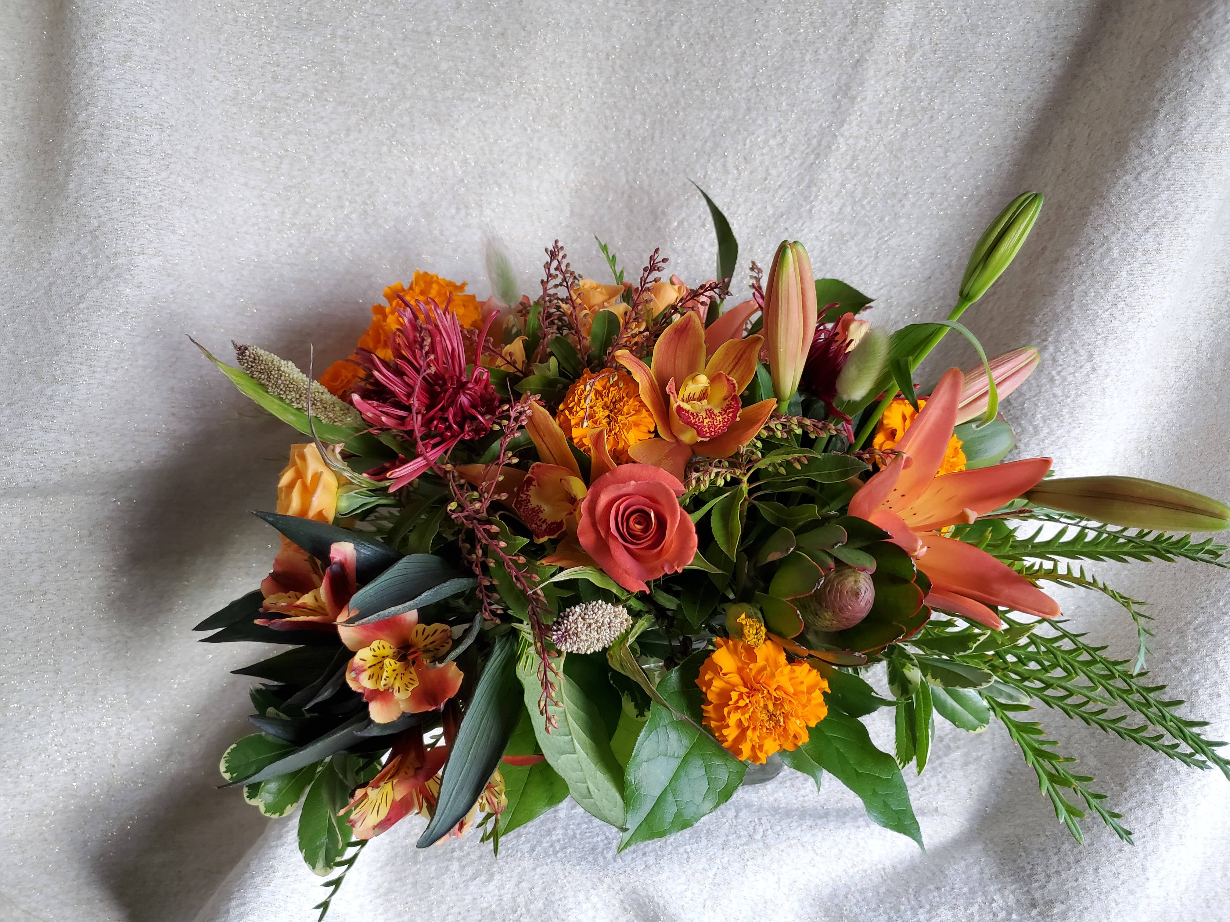 Low arrangement of orange lilies, roses, marigolds, orchids and mixed blooms in warm autumn tones