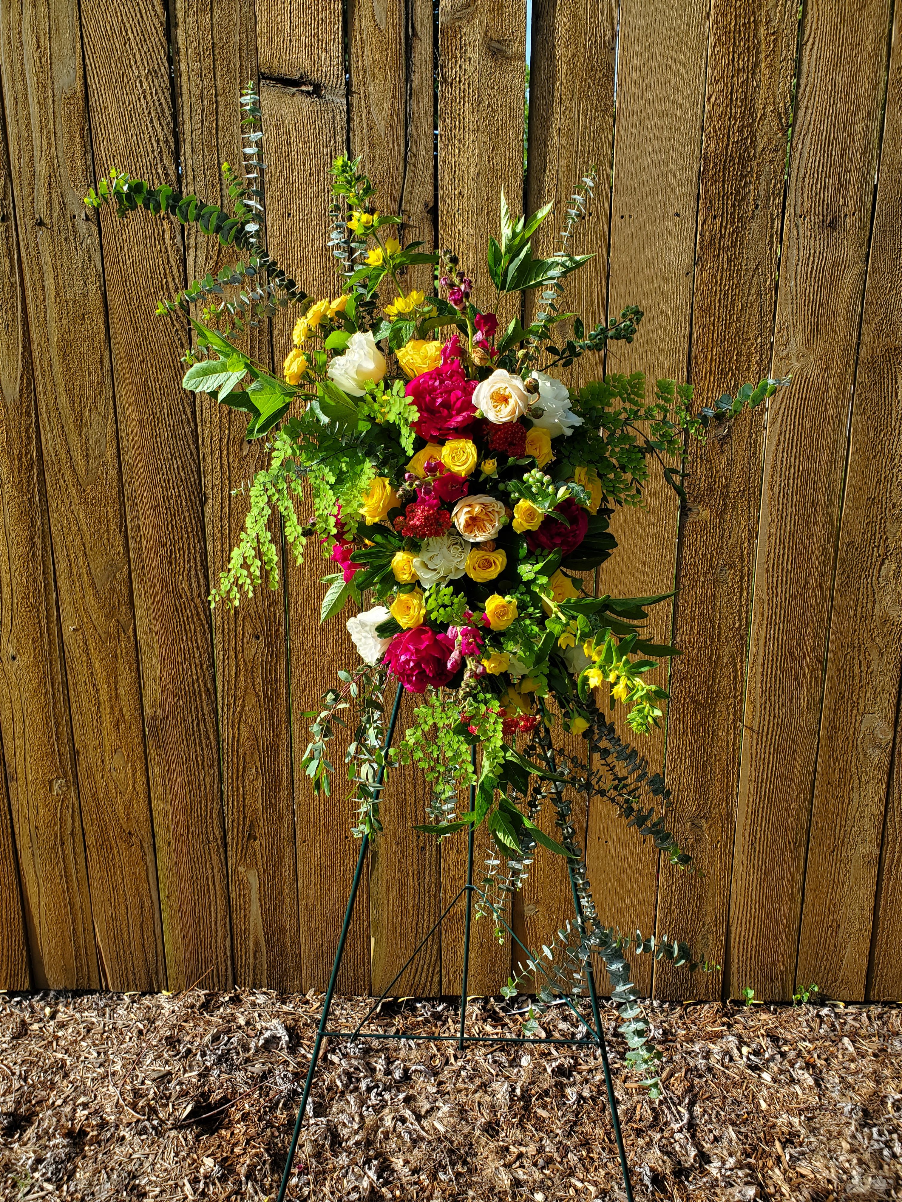 Bright standing spray of yellow, red, and white flowers on a metal easel against a wood fence.
