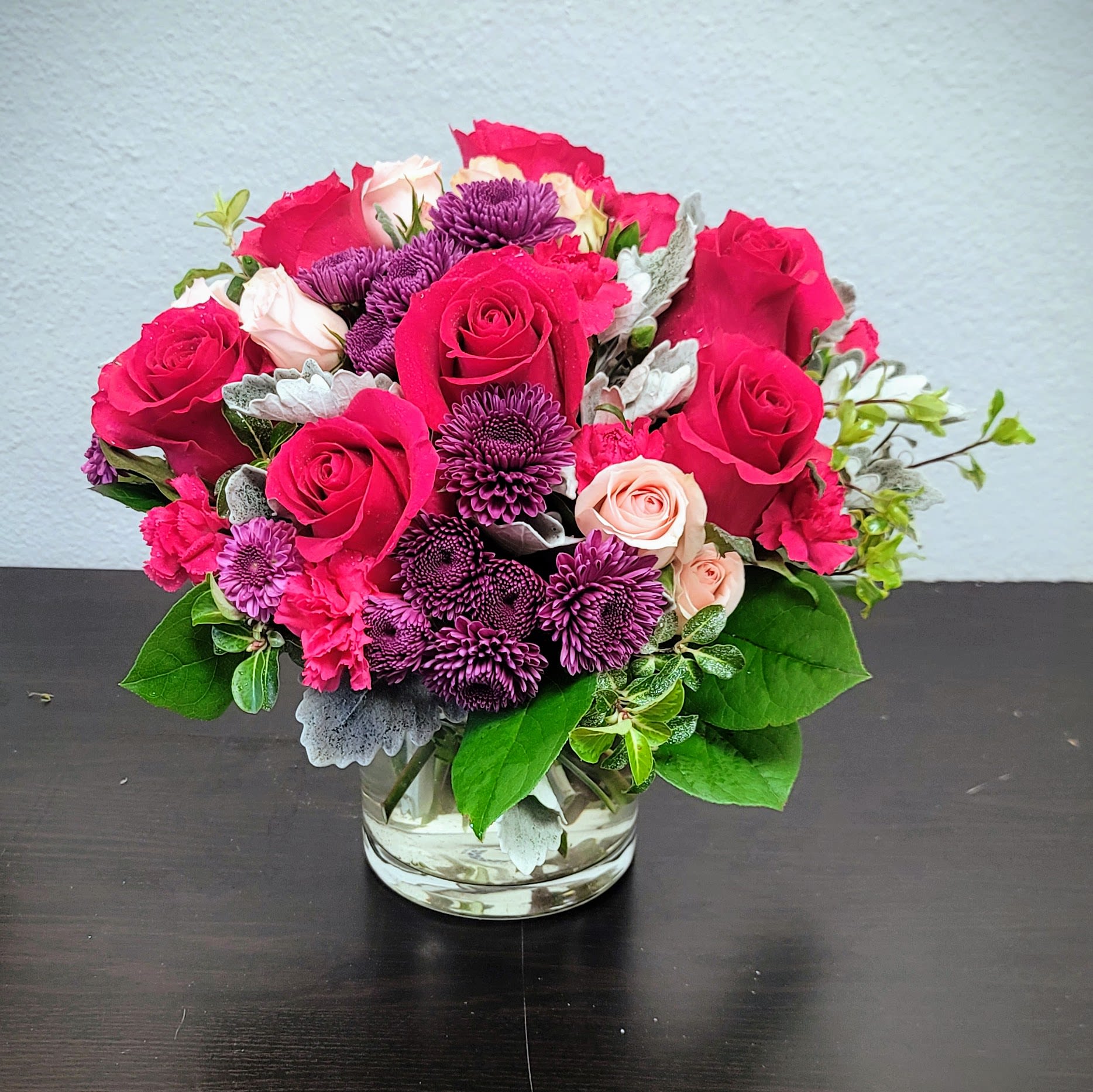Sweet As Can Be Bouquet - This stunning arrangement features deep red Roses, complemented by vibrant pink Carnations, and accented with deep purple Chrysanthemums.   The silvery foliage adds a touch of elegance and texture. Perfect for birthdays, anniversaries, Valentine's Day, or simply to express your love and admiration.  Size: Approximately 10" H and 12" W. Order now and brighten someone's day!