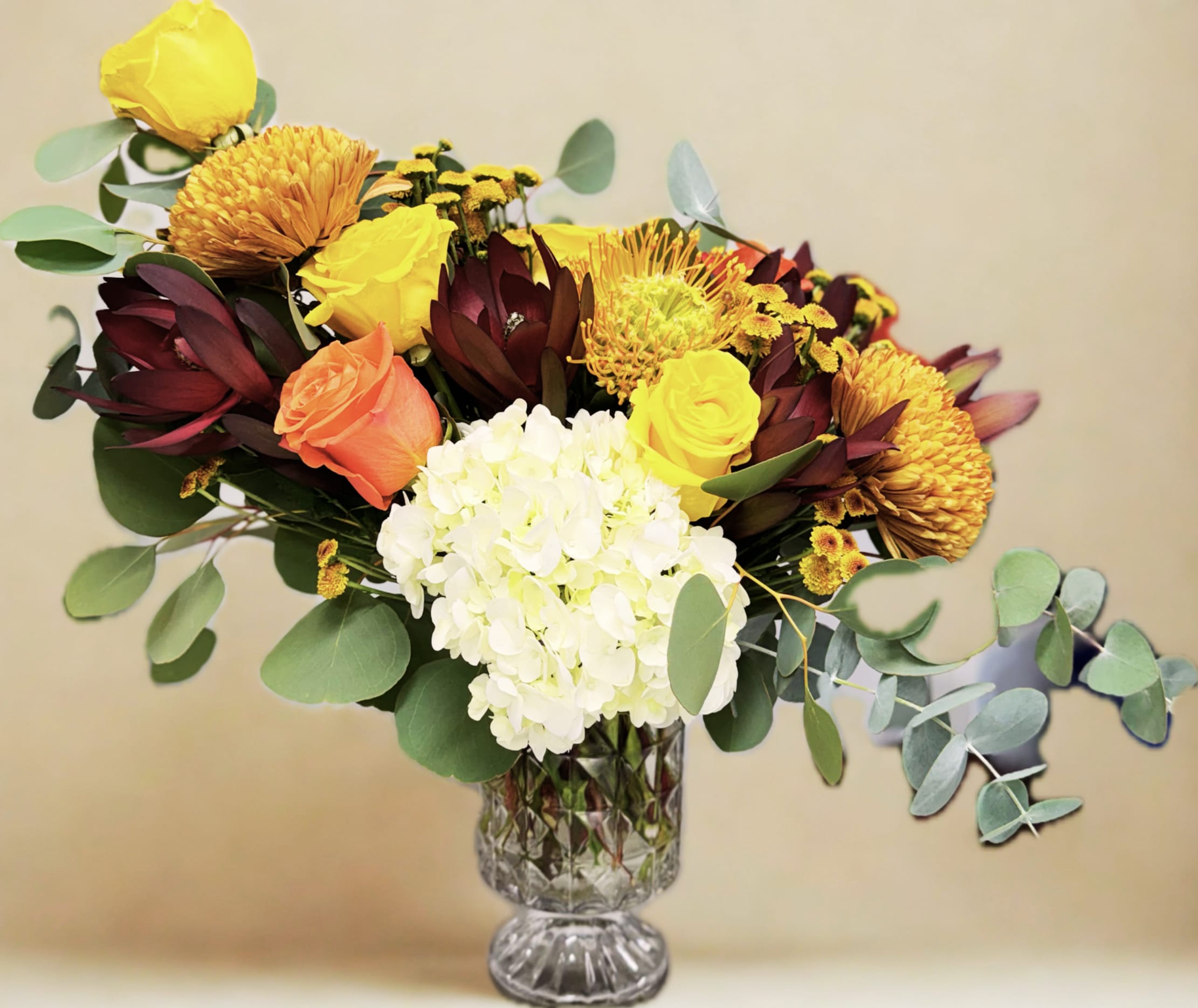 Jewels of fall floral arrangement  - Celebrate the richness of autumn with our Jewels of Fall arrangement — a vibrant blend of seasonal elegance and wild beauty. This lush bouquet features bold mums and velvety roses in deep, jewel-toned hues, accented by the exotic texture of pincushion protea and the striking foliage of Safari Sunset leucadendron. Arranged in a warm, natural palette of amber, rust, burgundy, and gold, it brings the perfect touch of autumn sophistication to any table or celebration.