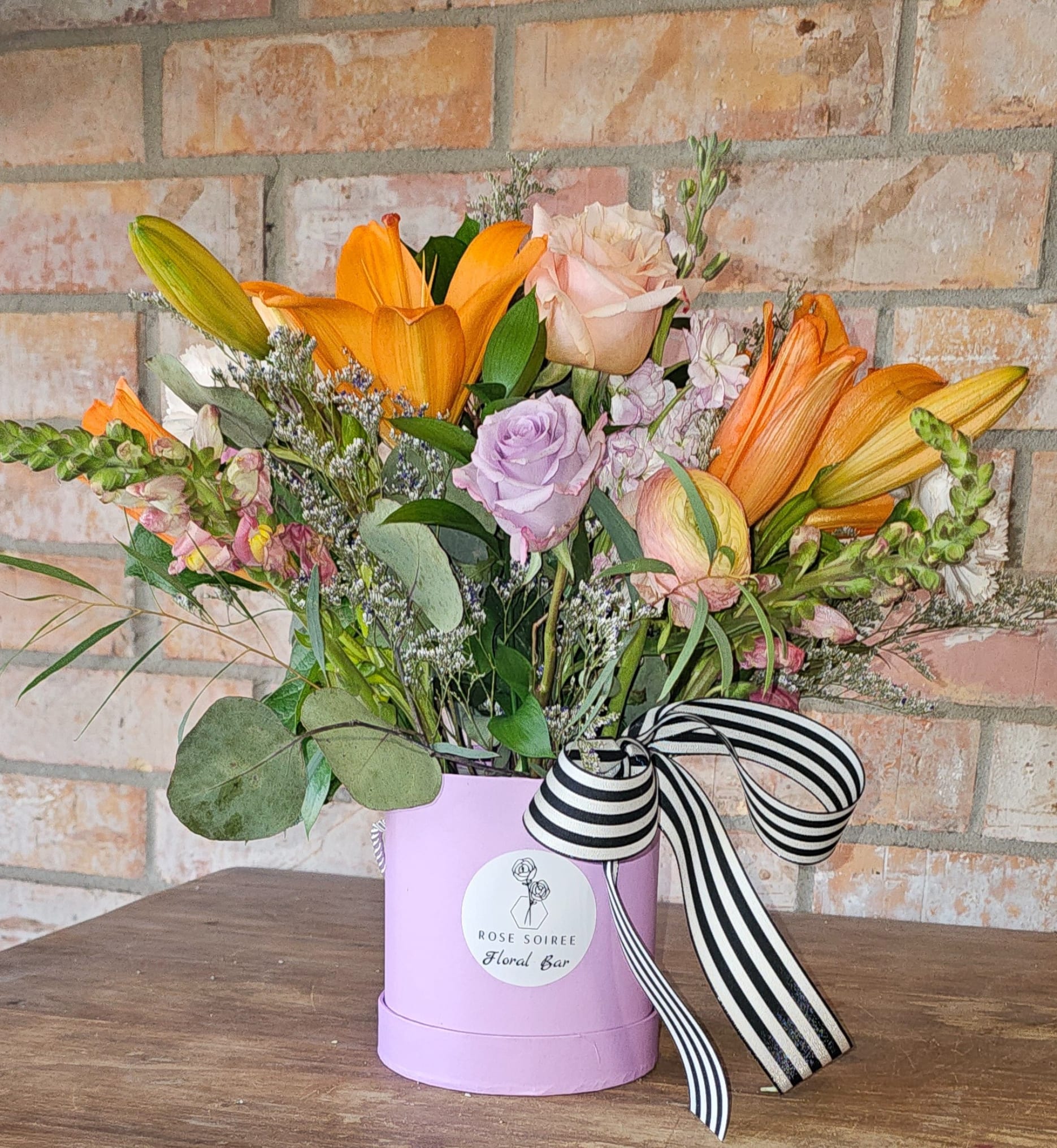 Mixed arrangement of orange lilies and pastel roses in a lavender hatbox with striped ribbon