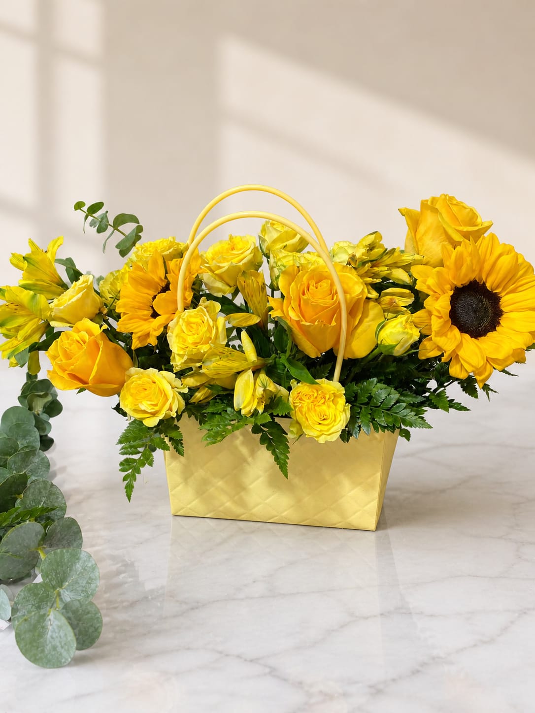 Flower bag yellow - Bright yellow roses and cheerful sunflowers spill from a gold textured basket with a curved handle - a sunny flower arrangement perfect for a birthday bouquet, thank-you gift, or spring celebration. The warm yellow and orange palette reads joyful and uplifting, the kind of floral gift that makes the person you're thinking of feel noticed and celebrated.  Tucked among the focal blooms are delicate yellow alstroemeria, eucalyptus sprigs, and fern-like foliage that add texture, contrast, and a fresh green backdrop. The piece arrives arranged in a gold basket with handle, giving it a polished presentation that's ready to display on a table or desk.  Ideal for letting someone know you care on an everyday occasion or marking a cheerful milestone, this compact arrangement offers long-lasting blooms and easy display - no extra vase required. Order with confidence for same-week gifting or to brighten a home or office with sunny, uplifting color.