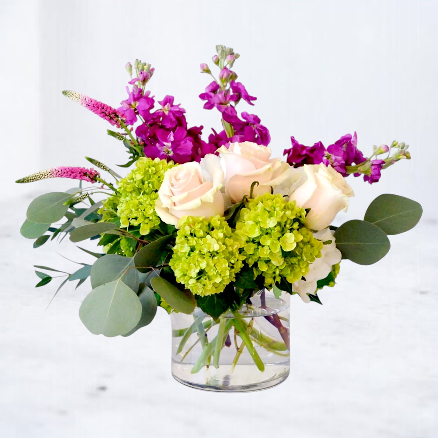 Weekender - Bring the carefree feeling of a perfect weekend home with Weekender, a vibrant garden-style flower arrangement designed to celebrate life's happiest moments. Featuring premium blush roses, vivid magenta stock, fresh green hydrangea, elegant veronica, eucalyptus, and a curated selection of seasonal blooms, this design is full of movement, texture, and joyful color.   Handcrafted in a modern glass cylinder vase, Weekender blends soft peach tones with lively pinks and fresh greens to create an arrangement that's both playful and refined. The naturally gathered style gives every bloom room to shine, resulting in a fresh-from-the-garden look that's equally at home on a dining table, kitchen island, office desk, or entryway.  Whether you're celebrating a birthday, congratulating a friend, welcoming a new baby, sending thanks, or surprising someone "just because," Weekender makes every occasion feel a little brighter. Its cheerful palette and contemporary design make it a thoughtful gift that brings instant warmth and lasting smiles.