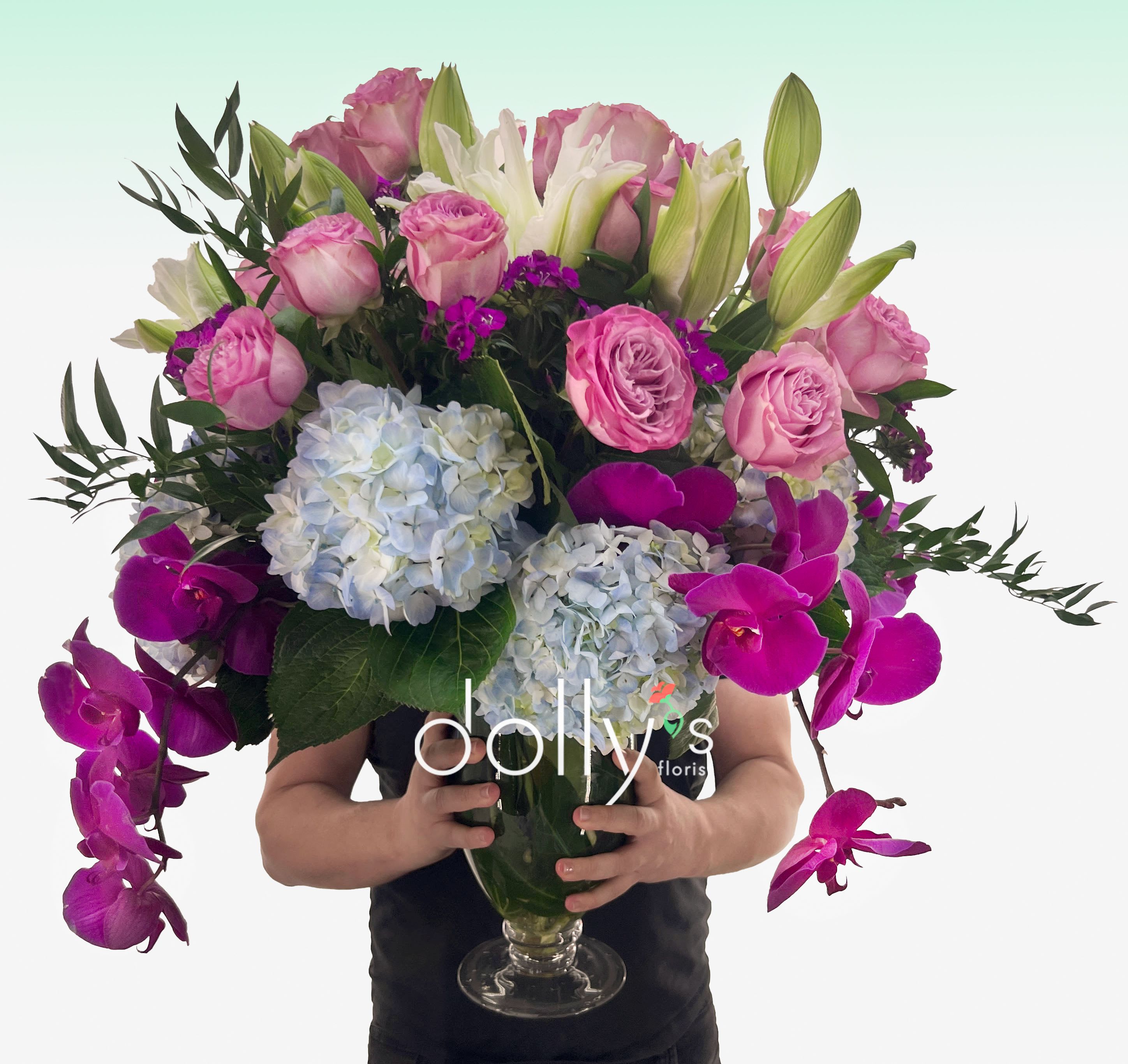 Tall arrangement of pink roses, white lilies, blue hydrangeas, and fuchsia orchids in a clear glass vase