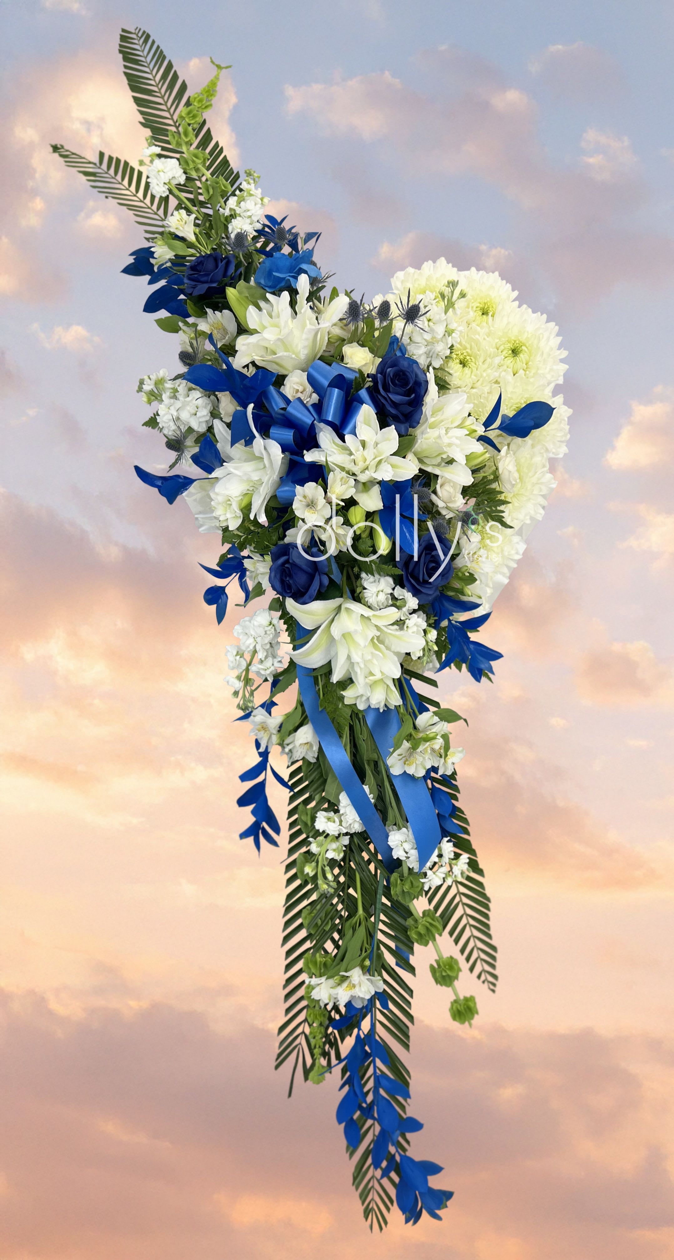 Blue | Bleeding Heart  - A tall, cascading sympathy spray that reads with ceremonial grace: white lilies and chrysanthemums ripple down beside deep blue roses and blue accent blooms, the composition falling in an elegant vertical line that gently guides the eye. Designed for memorials and funeral services, this standing arrangement offers a respectful, solemn silhouette-palm-like fronds and trailing blue ribbon add movement and a dignified finish so the person you're honoring feels truly remembered.  Measuring approximately 24 x 22 inches (60.96 cm x 55.88 cm), the arrangement pairs crisp white blooms with striking blue roses and touches of green foliage for a clean, formal contrast. The asymmetrical cascade is intended for display on a stand and includes blue ribbon among the hard goods for a cohesive presentation. Specify preferred color notes in the special instructions box when ordering.