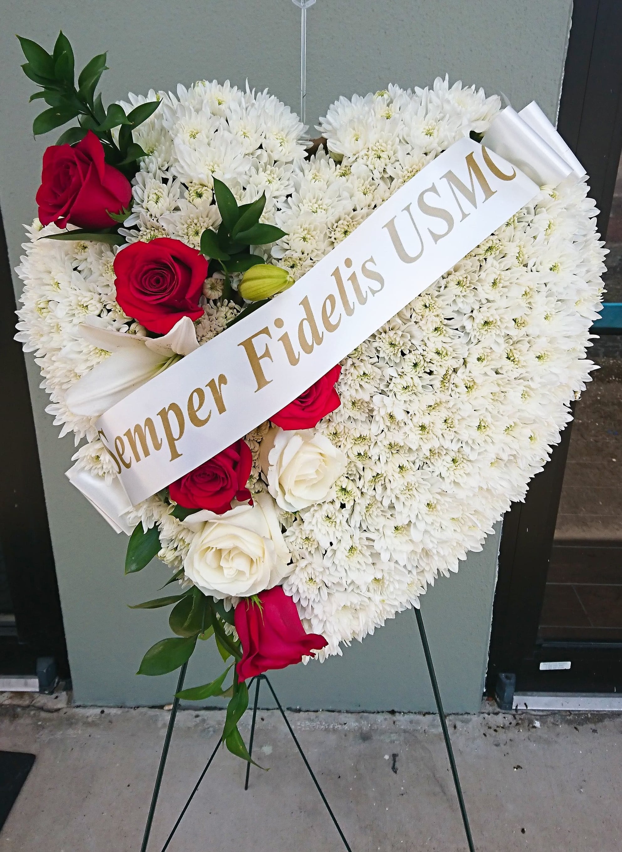 Salute to a Patriot Standing Heart - TheD Salute to a Patriot Standing Heart is the perfect tribute  to a fallen marine. A pearly white heart consisting of chrysanthemums is accented with an arrangement of red roses, white Asiatic lilies and lush greens, as well as, a white banner that reads: "Semper Fidelis USMC" to create an incredible presentation at their final memorial service.   Displayed on a wire easel. Dimensions are 24"h x 24"w.