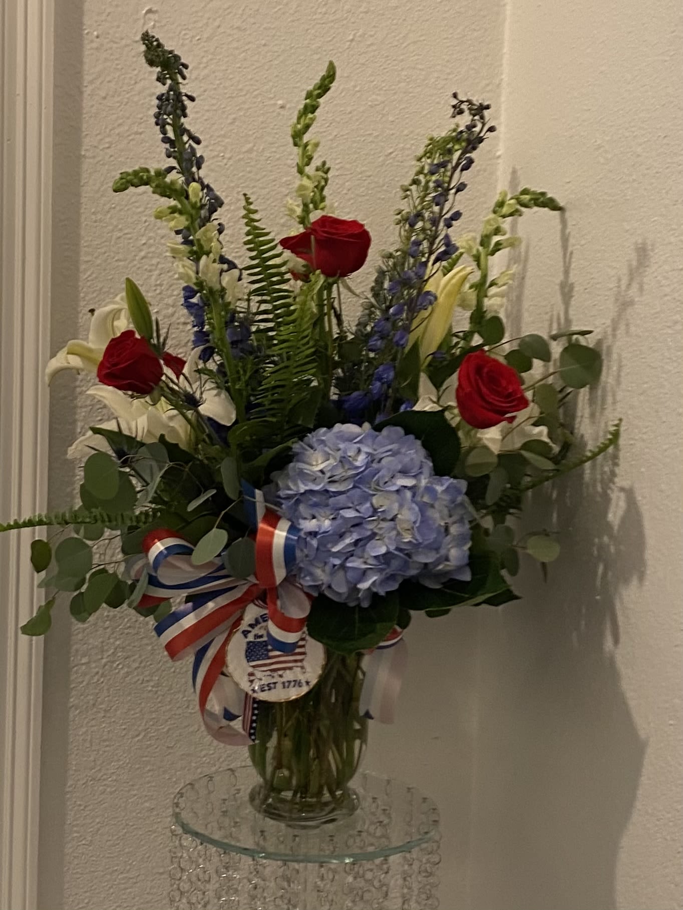 Red, White and Boom - A bold red, white and blue arrangement that reads like a salute-perfect as a patriotic centerpiece, Fourth of July gift, or celebratory bouquet for graduations and military milestones. This tall, upright flower arrangement places large blue hydrangea blooms at the heart, framed by velvety red roses and fragrant white lilies so the person you're thinking of will feel honored and noticed the moment it arrives.  Slender blue delphinium spikes and airy greenery give the bouquet height and movement, while the clear glass vase keeps the look crisp and elegant. A red, white and blue ribbon and a round patriotic decoupaged shell finish the presentation for a distinctly American theme. The mix of textures-from soft hydrangea petals to structured rose heads and the lilies' trumpet shapes-creates visual depth and a festive, refined feel.  Ideal for patriotic holidays, congratulations, or to send appreciation to someone who loves traditional colors, this arrangement arrives ready to display and makes a striking statement on a mantel, dining table, or office desk. Follow standard floral care-fresh water in the vase and trimming stems as needed-to extend the life of the blooms.