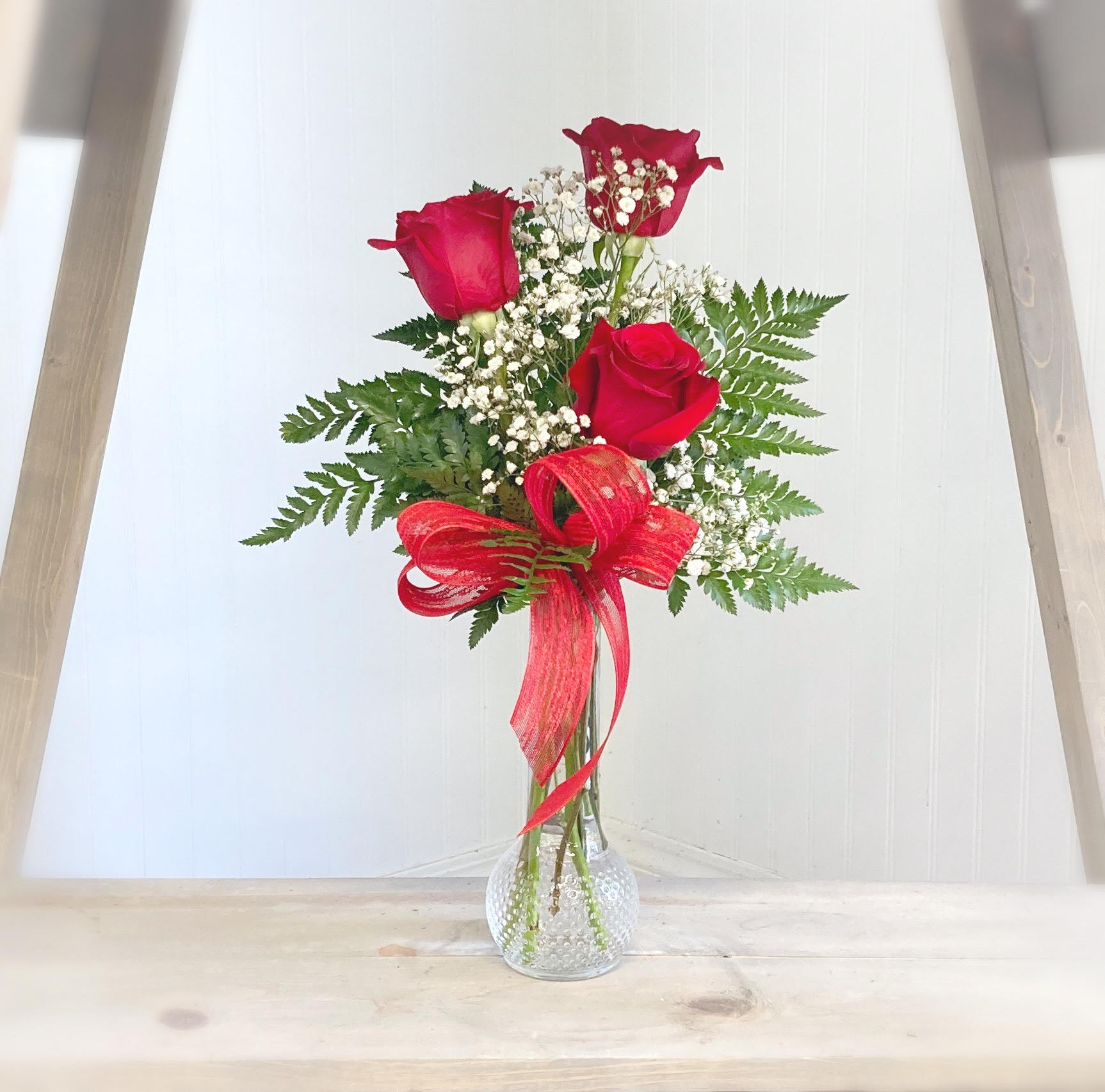 Three red roses in a clear glass vase with baby's breath and a red ribbon