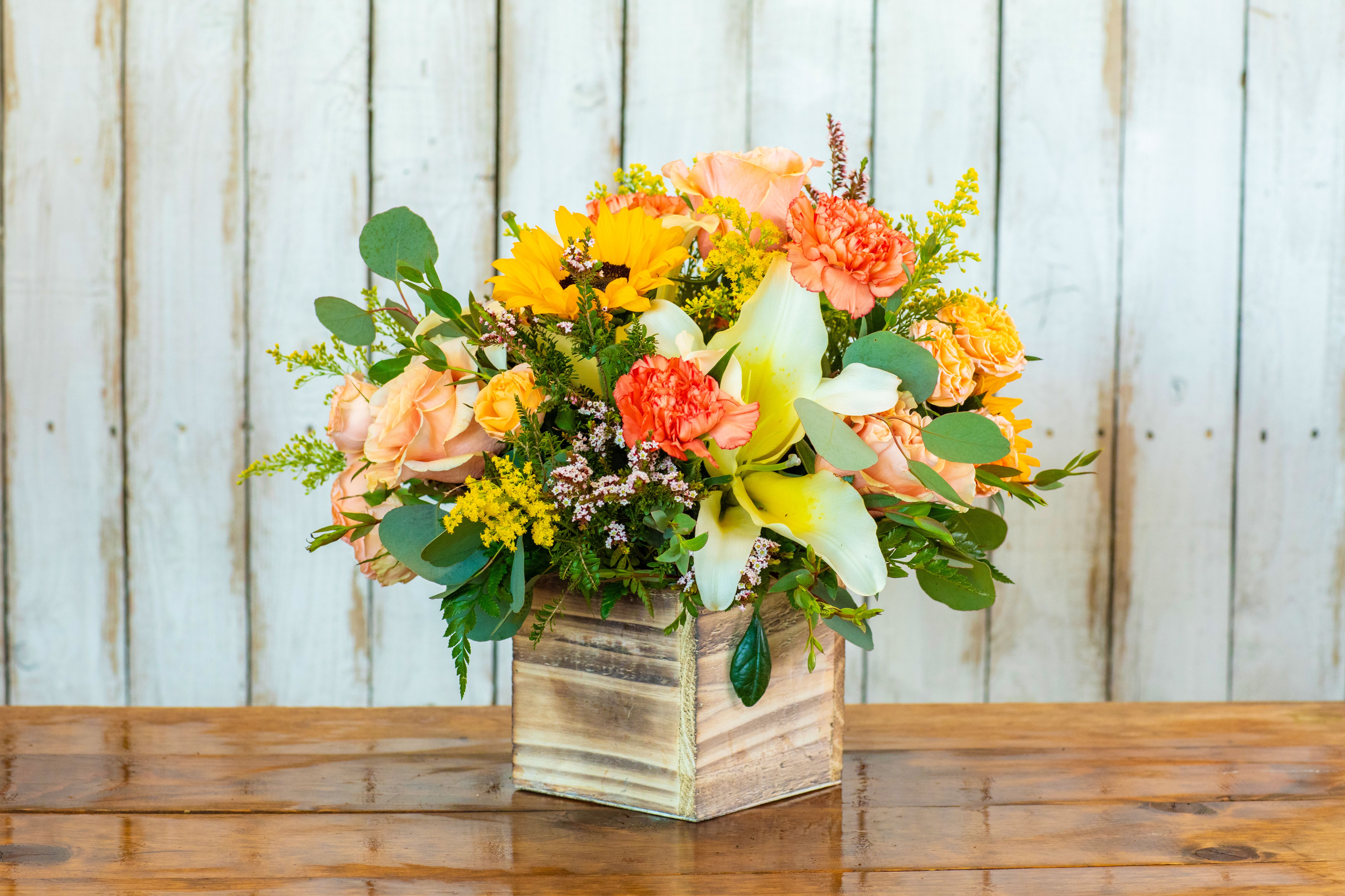 Rancho Sunset Flower Box - Capture the warm glow of a California evening with the Rancho Sunset Flower Box, a vibrant and radiant arrangement from your favorite Rancho Cucamonga Florist. This lively cube arrangement is a feast for the eyes, featuring a cheerful mix of sunny colors and textures.  The design centers around brilliant sunflowers and large, creamy white lilies, surrounded by ruffled peach roses and bi-color orange carnations. Accents of golden solidago (filler flowers) and silvery eucalyptus leaves are nestled inside a charming, rustic wooden box, which provides a lovely, natural container.  As a leading Rancho Cucamonga flower shop, we are experts in designing high-quality arrangements that convey happiness and warmth. This sunny bouquet is the perfect choice for a joyful birthday bouquet, a meaningful anniversary gift, a thoughtful get well soon surprise, or a housewarming present. It's especially wonderful for celebrating autumn or sending a burst of summer cheer!  We guarantee the freshest blooms and offer reliable same-day flower delivery throughout the Rancho Cucamonga area. Trust our dedication to quality and service. Order the Rancho Sunset Flower Box online today from your dependable local Rancho Cucamonga Florist!