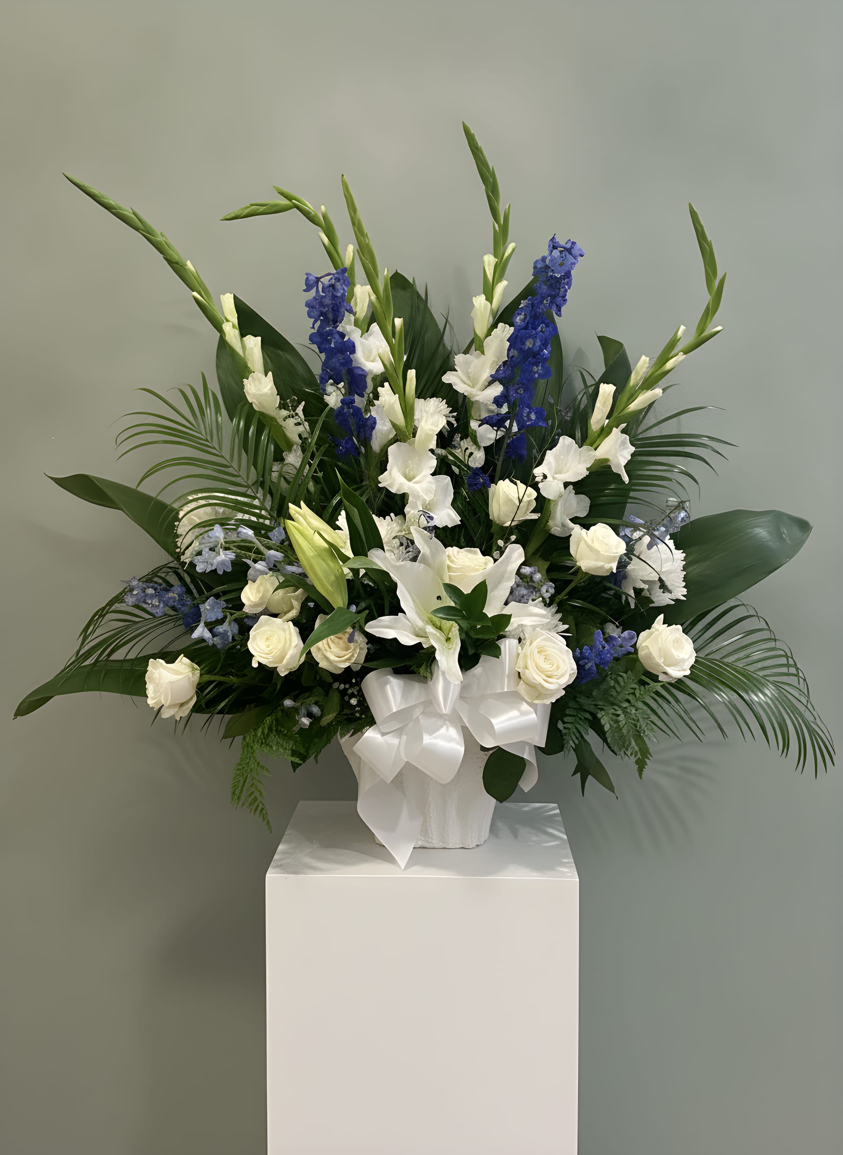 Heavenly Blue &amp; White Tribute Basket - Honor a loved one with our elegant Heavenly Blue &amp; White Tribute Basket, a timeless sympathy arrangement designed to express love, remembrance, and peace. This beautiful standing basket features premium white lilies, white roses, gladiolus, blue delphinium, carnations, and seasonal accent flowers surrounded by lush greenery.  The classic combination of blue and white flowers symbolizes faith, serenity, and everlasting love, making this arrangement a thoughtful choice for funeral services, memorial celebrations, church ceremonies, and expressions of sympathy.  Handcrafted by the designers at Tommy Austin Florist, each tribute basket is created with fresh, high-quality flowers and professional attention to detail.     Premium white lilies and roses * Blue delphinium accents * White gladiolus and carnations * Fresh seasonal flowers and greenery * Suitable for funerals, memorial services, and celebrations of life * Hand-delivered by Tommy Austin Florist