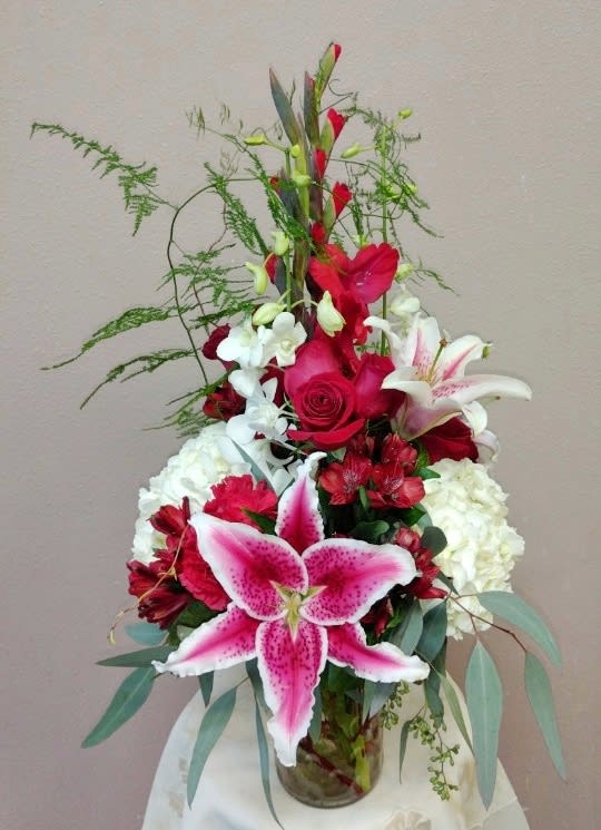 Tall red, white, and pink flower arrangement with lilies, roses, orchids, and hydrangeas in a clear glass vase.