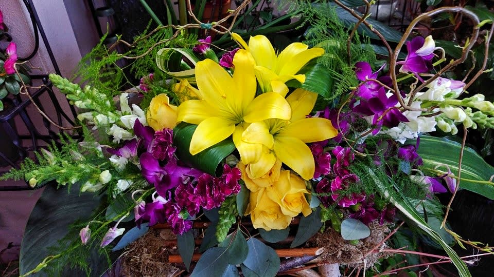 Bright yellow lilies and purple orchids arranged with roses in a decorative basket.