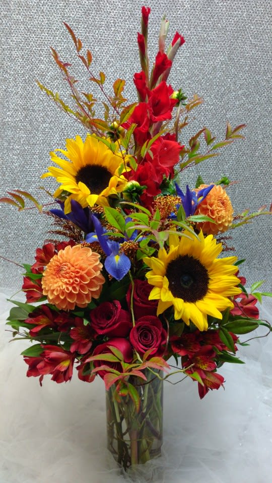 Tall mixed arrangement with sunflowers, red roses, orange dahlias, and purple iris in a clear glass vase