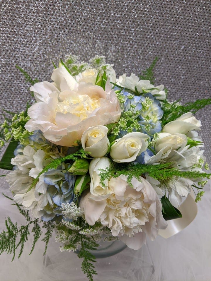 Round bouquet of ivory peonies, white roses, and pale blue flowers in a glass vase.