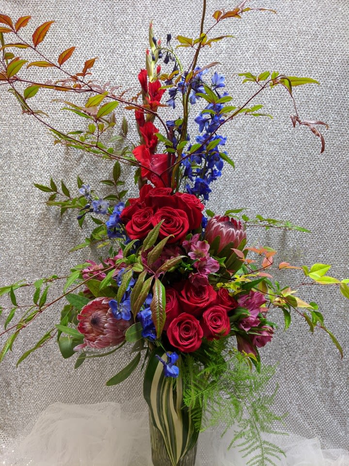 Tall bouquet of red roses, blue flowers, and protea in a glass vase