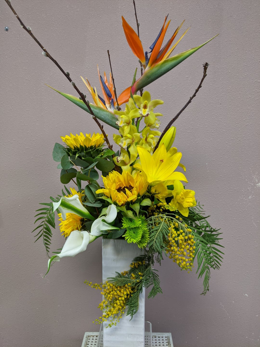 Tall floral arrangement with yellow blooms, white calla lilies, and bird of paradise in a white vase