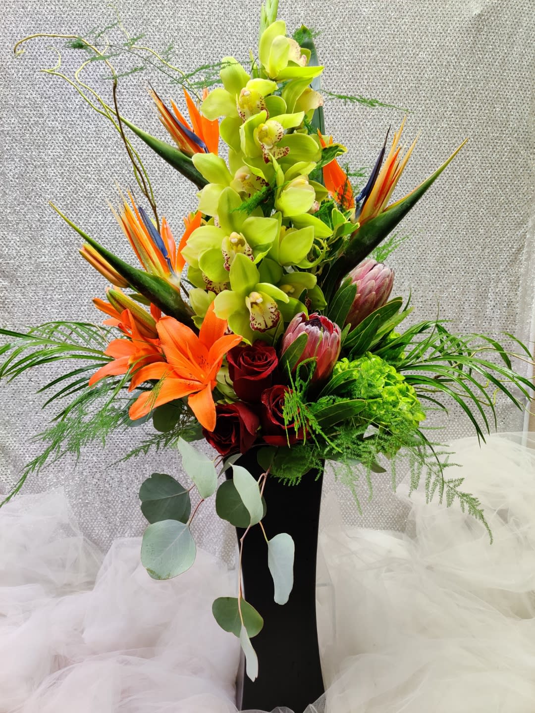 Tall tropical bouquet with green orchids, orange lilies, and red roses in a black vase