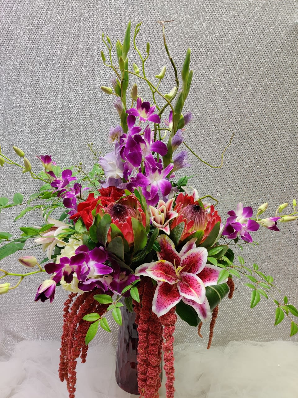 Tall tropical arrangement with purple orchids and pink lilies in a dark vase