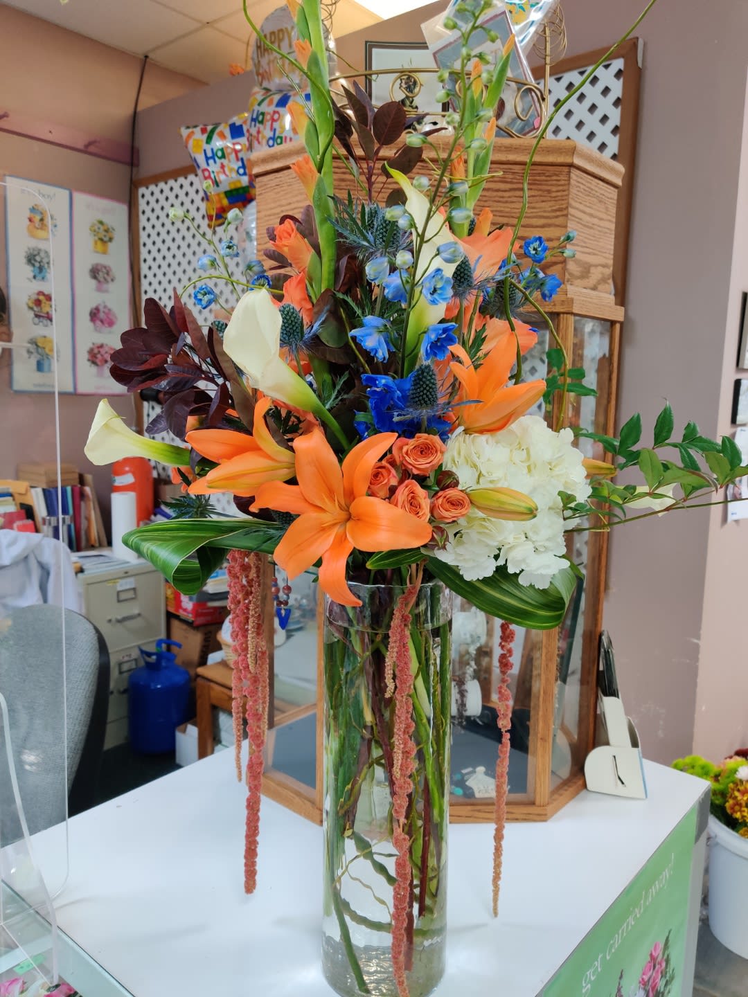 Tall mixed bouquet with orange lilies, blue accents, and white blooms in a glass vase