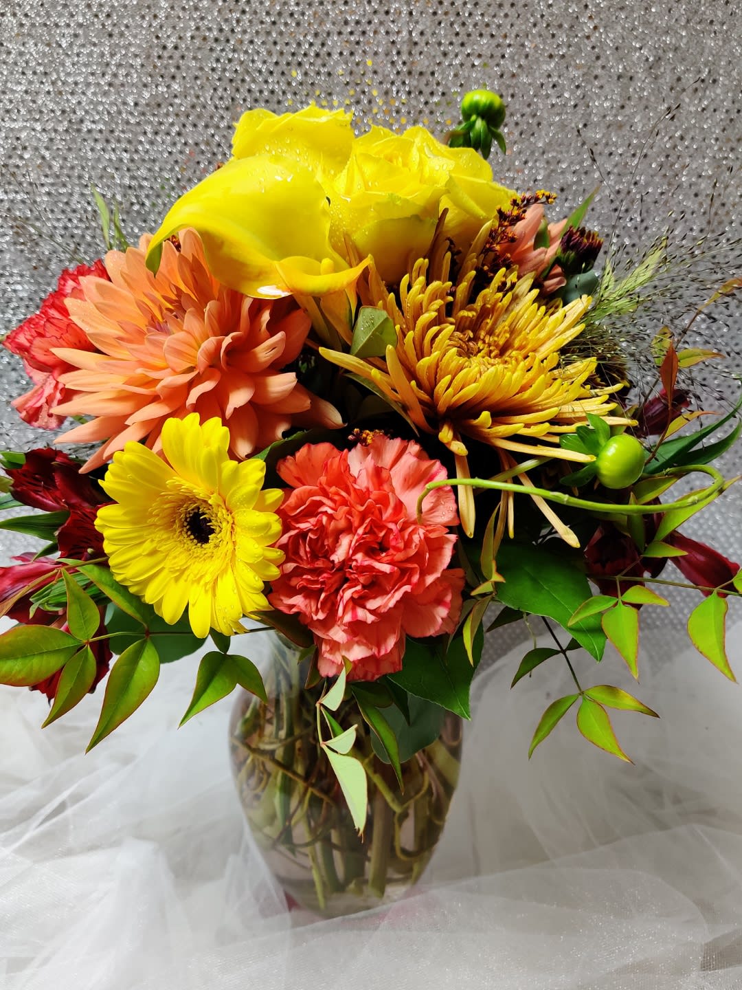 Bright mixed bouquet in a clear glass vase with yellow, peach, and coral blooms.