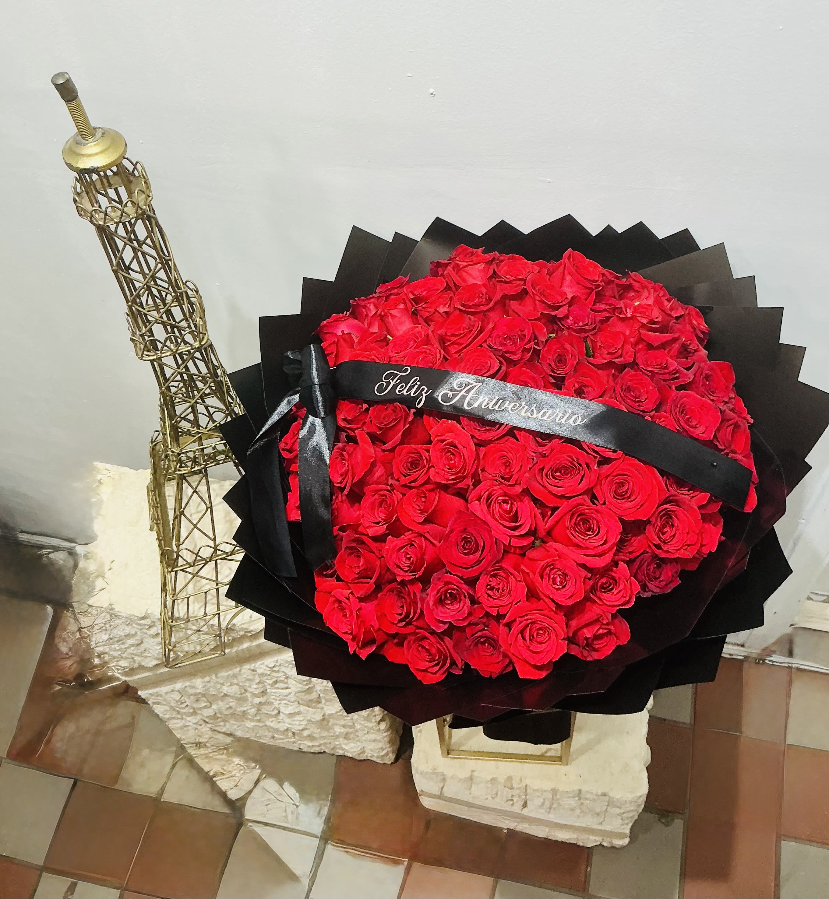 Feliz aniversario - A dense, hand-tied bouquet of vivid red roses makes an immediate, romantic statement - the perfect anniversary bouquet to tell someone "Feliz aniversario" in a way that feels both dramatic and intimate. Arranged into a rounded, almost heart-like mound, these red roses wrapped in layers of matte black paper and tied with a black satin ribbon printed with an anniversary message deliver a bold contrast that highlights the velvet texture and deep color of each bloom. This design also works as a meaningful "I'm sorry" or love-and-romance gesture that reads clearly from across the room.  The arrangement is offered two stylish presentations: a tall display on a slim metal stand with a wire Eiffel Tower sculpture for a striking, gallery-style moment, or a low, hatbox-style presentation framed by angular black wrap and a gold-toned metal tower sculpture for a luxe tabletop gift. Finished with the printed black ribbon and set in a round hatbox-style container, the piece measures as a substantial, show-stopping focal arrangement ideal for anniversaries, spring celebrations, or Día de las Flores Amarillas. Delivered in Saint Paul, MN, it's designed to make the person you're thinking of feel celebrated, forgiven, or deeply loved - order with confidence for same-day options when available.