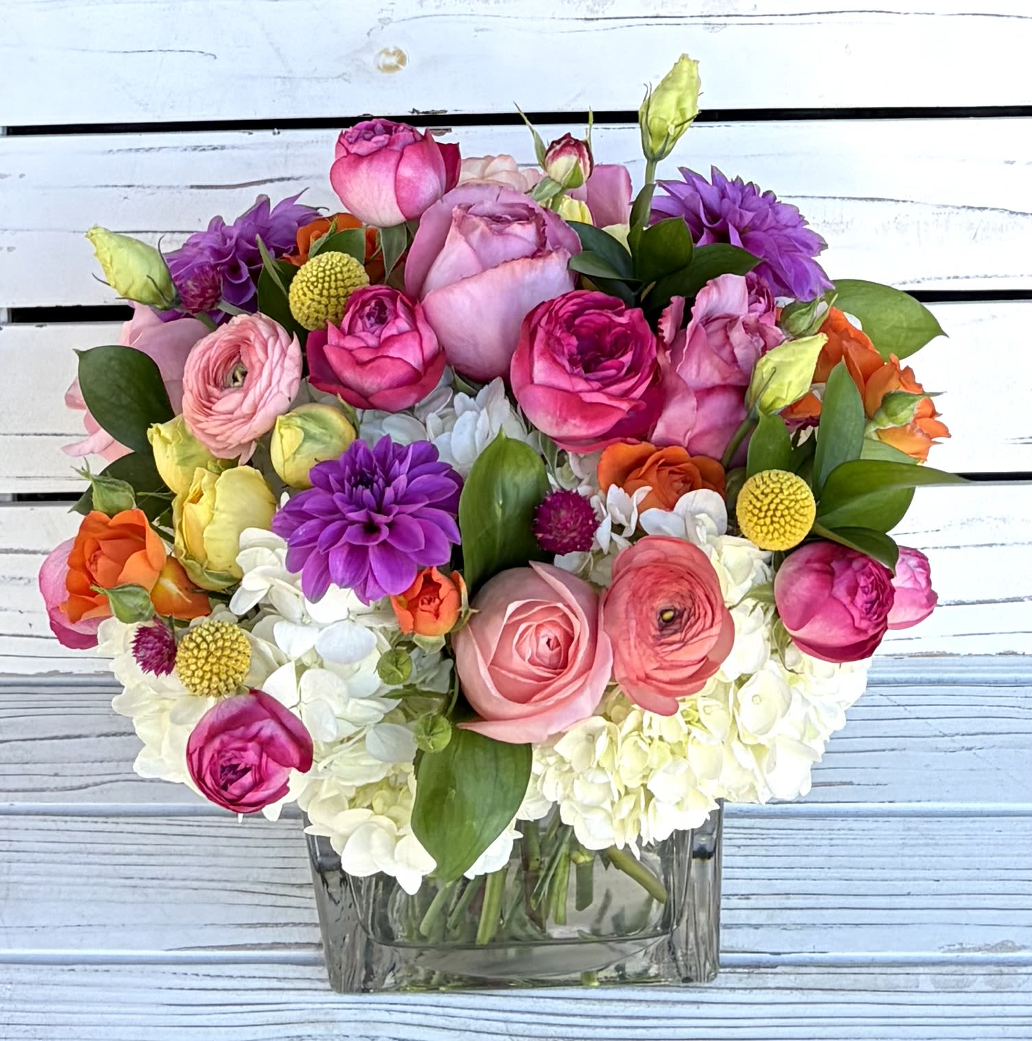 Colorful arrangement of pink roses, ranunculus, dahlias and white hydrangeas in a clear glass cube vase.