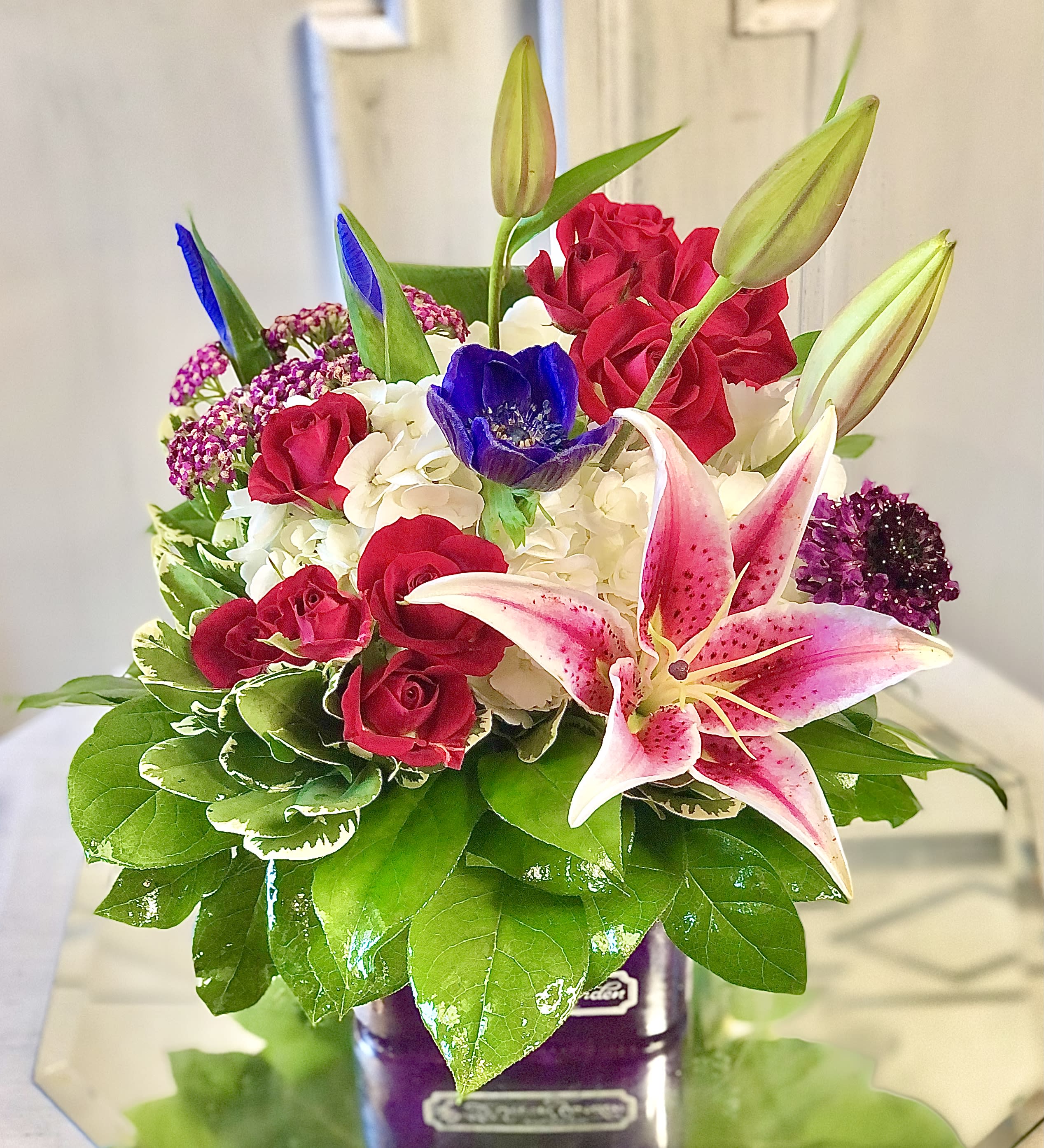 Midnight Magic - Dark and rich jewel tone colors create a magical design in this featured design, Midnight Magic. Includes lilies, garden roses, iris, anemone, and other garden flowers. This arrangement is approximately 11.5in in height and 9.5in in width.