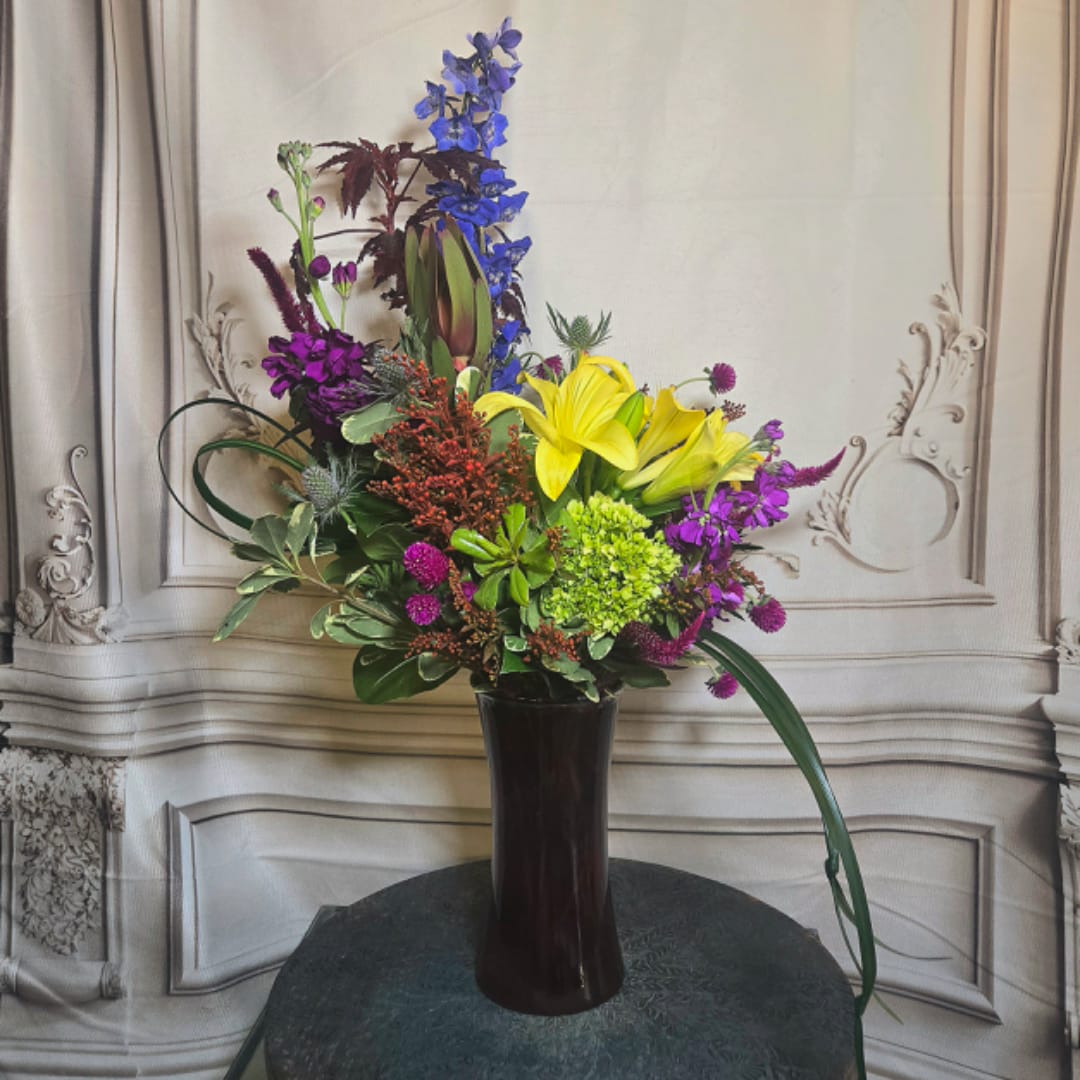 Tall mixed flower arrangement in a dark vase with yellow lilies and purple blooms