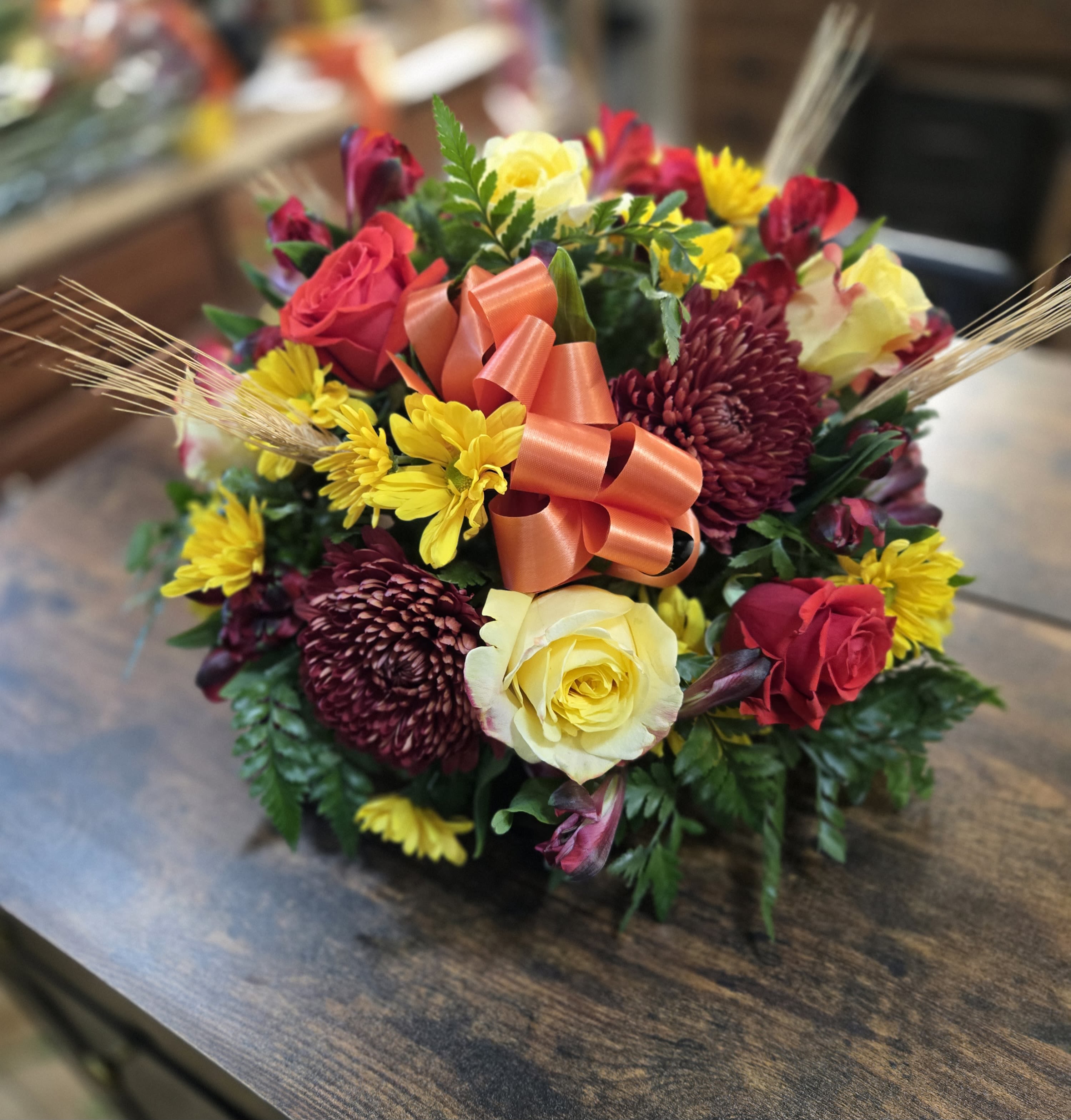 Autumn harvest centerpiece  -  It’s a full, lush fall-themed arrangement with roses, yellow spray mums, burgundy mums, alstroemeria, greenery, wheat accents, and a large decorative orange bow
