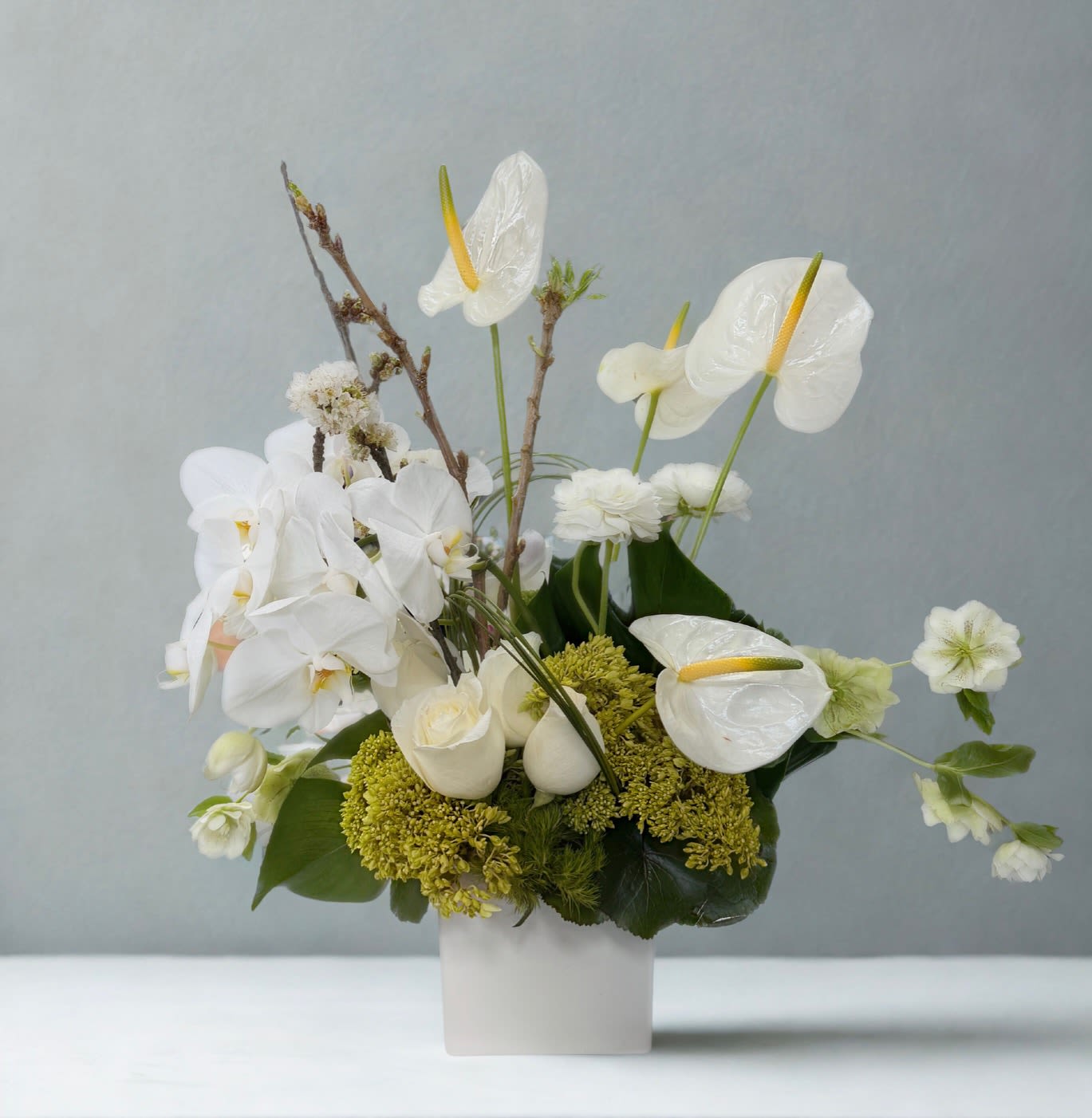Graceful White - A modern, sculptural arrangement in soft ivory and fresh green tones, designed to feel elegant, serene, and effortlessly refined. This piece features white phalaenopsis orchids, glossy white anthuriums, creamy roses, delicate seasonal blooms, and textured green accents arranged in a sleek white vessel. With its clean palette and contemporary silhouette, it makes a beautiful choice for sympathy sentiments, sophisticated home décor, or any occasion that calls for understated luxury.  