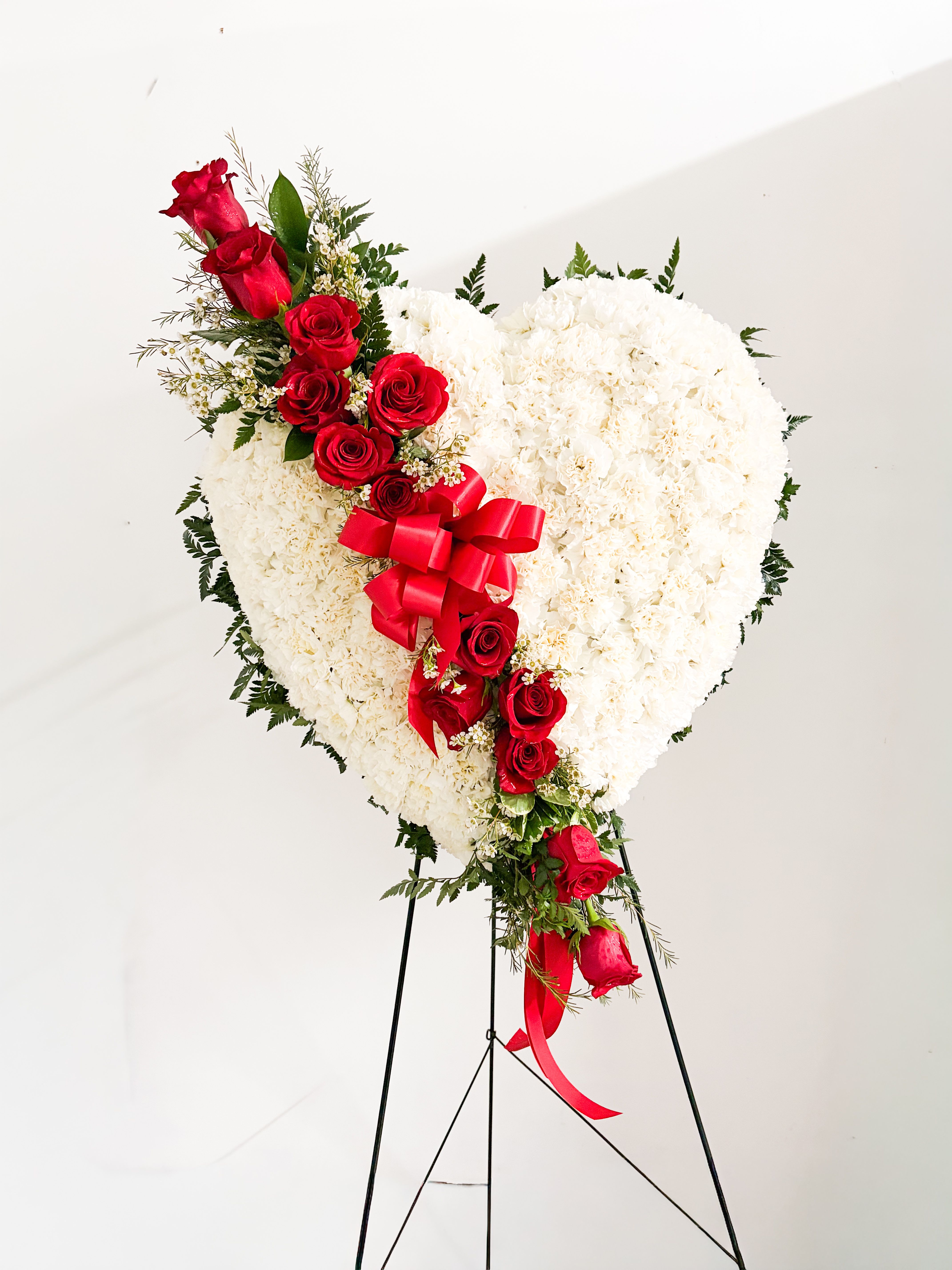 LOVE HEART STANDING SPRAY - A large heart-shaped standing spray that speaks to the person you're honoring with a clear message of love and remembrance: this sympathy and funeral arrangement combines red roses and white carnations in a formal, heart-shaped tribute perfect for thinking of you moments. The diagonal cascade of deep red roses across a cushion of tightly packed white carnations creates a striking red-and-white contrast that reads both romantic and reverent, so the recipient feels truly seen and remembered.  Made on a heart-shaped wreath form and finished on a metal easel stand, the spray is accented with mixed greenery, small white filler blooms, and a bright red satin ribbon bow with trailing tails for a dignified focal point. The dense, cushion-like white carnations provide a full, lasting base while the red roses add texture, depth, and emotional clarity. Ideal for memorial services, graveside display, or sending sympathy and thinking-of-you condolences , this arrangement arrives ready to display and offer comfort.