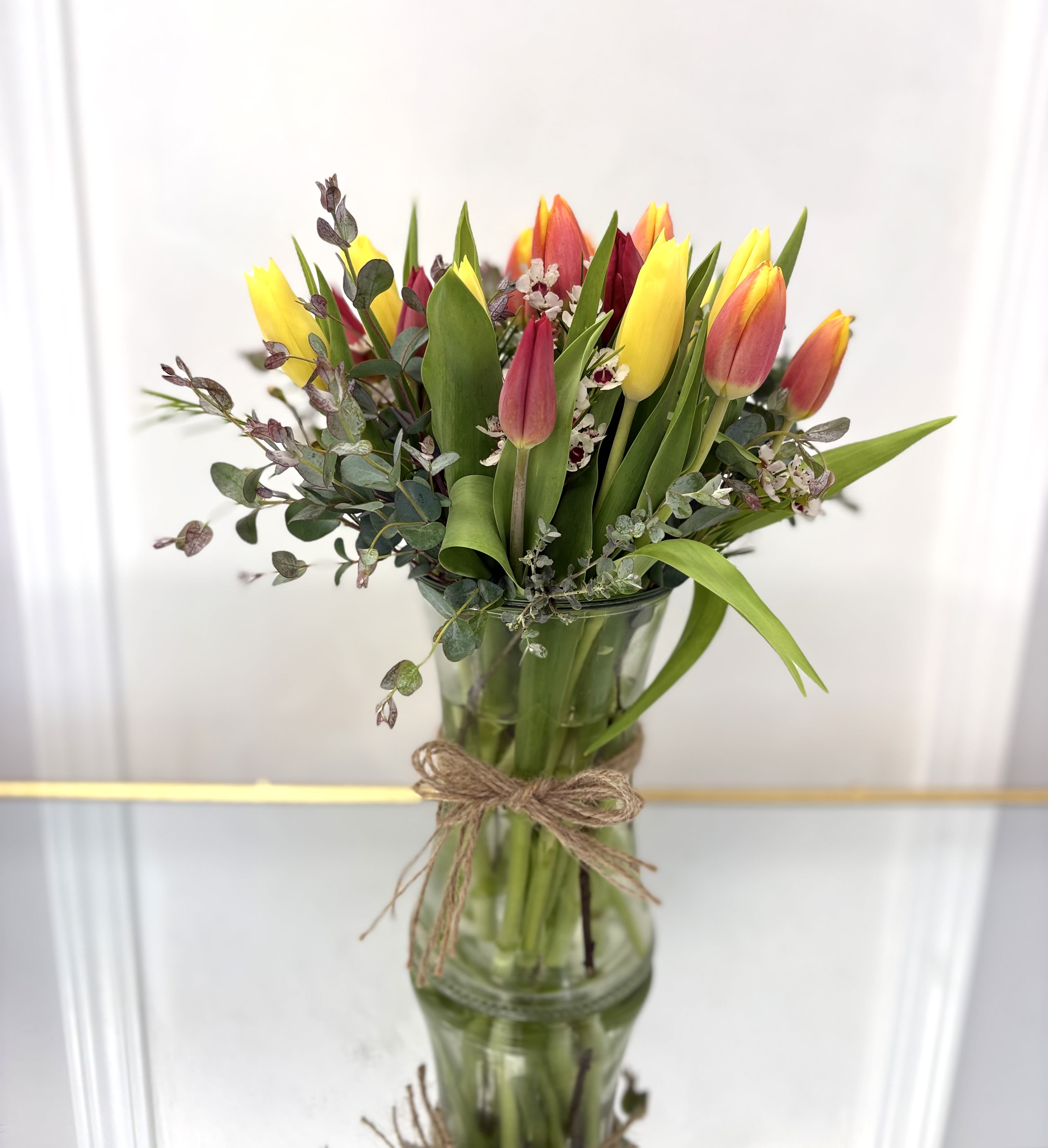 Yellow and red tulips with small white blooms in a tall clear glass vase tied with twine
