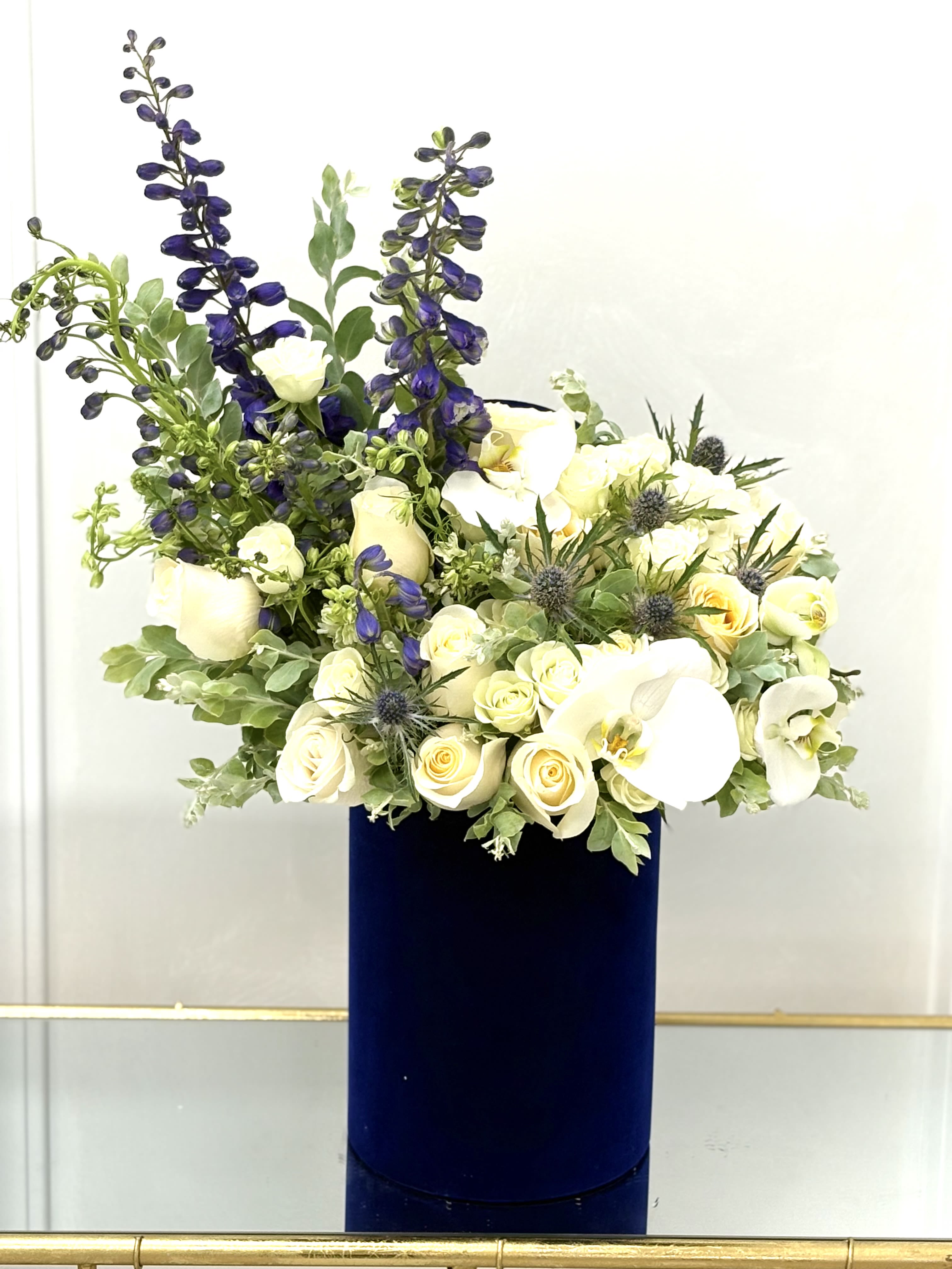 White roses and orchids in a tall navy vase with blue delphinium spikes