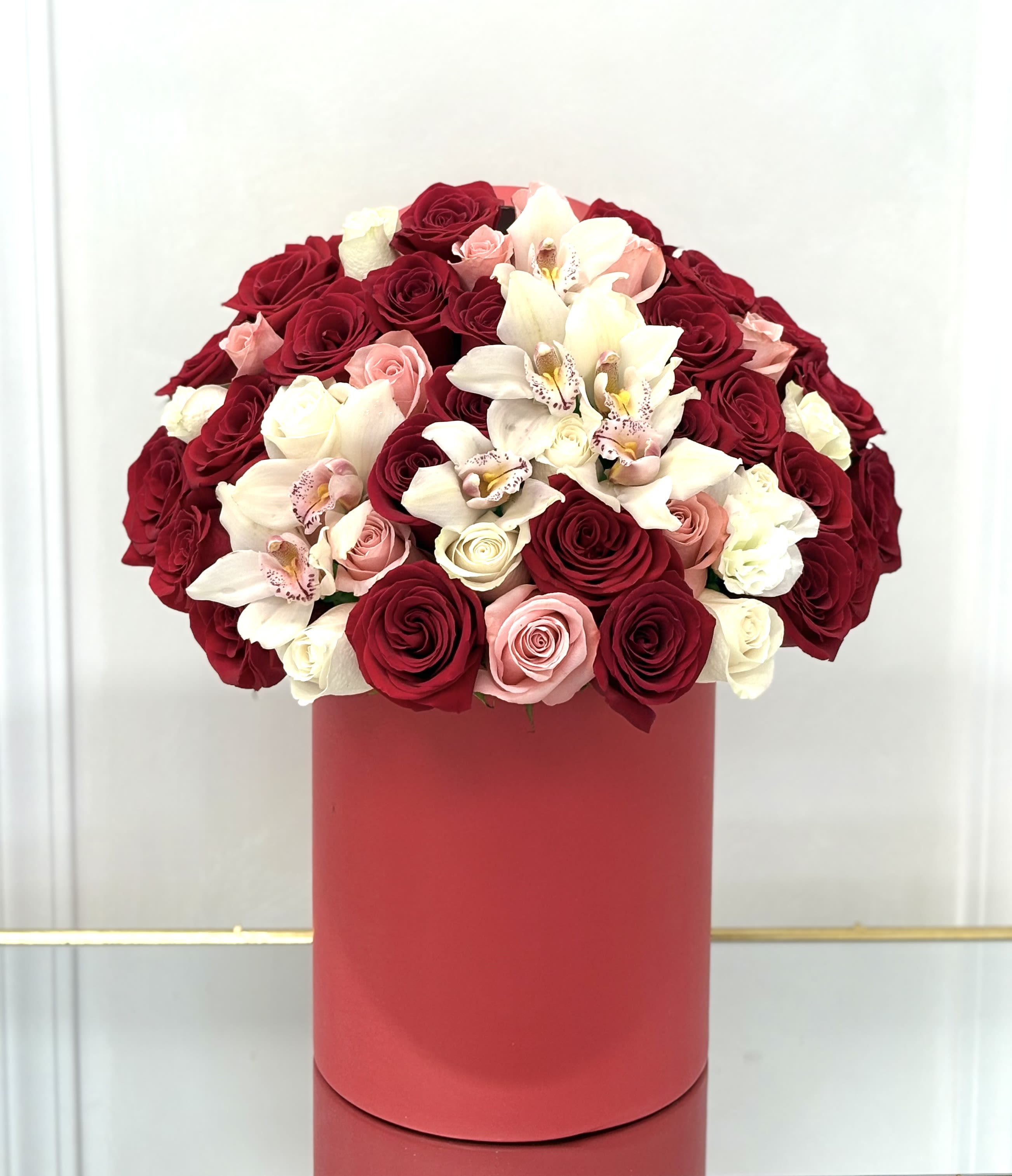Round arrangement of red, pink, and white roses with orchids in a tall red hatbox-style container