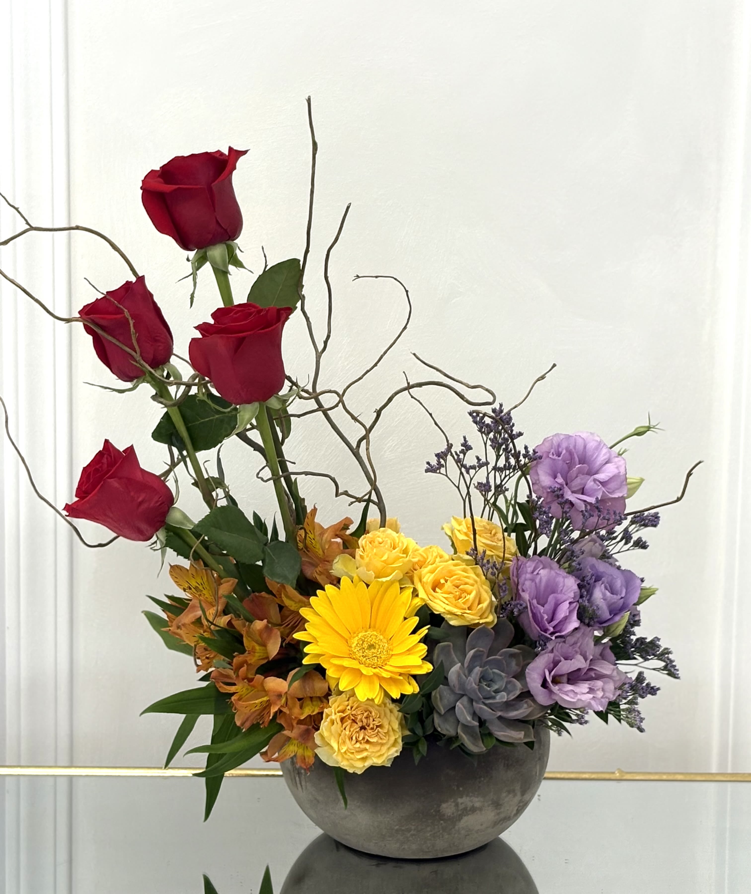 Mixed floral arrangement with red roses, yellow blooms, and purple flowers in a gray bowl vase