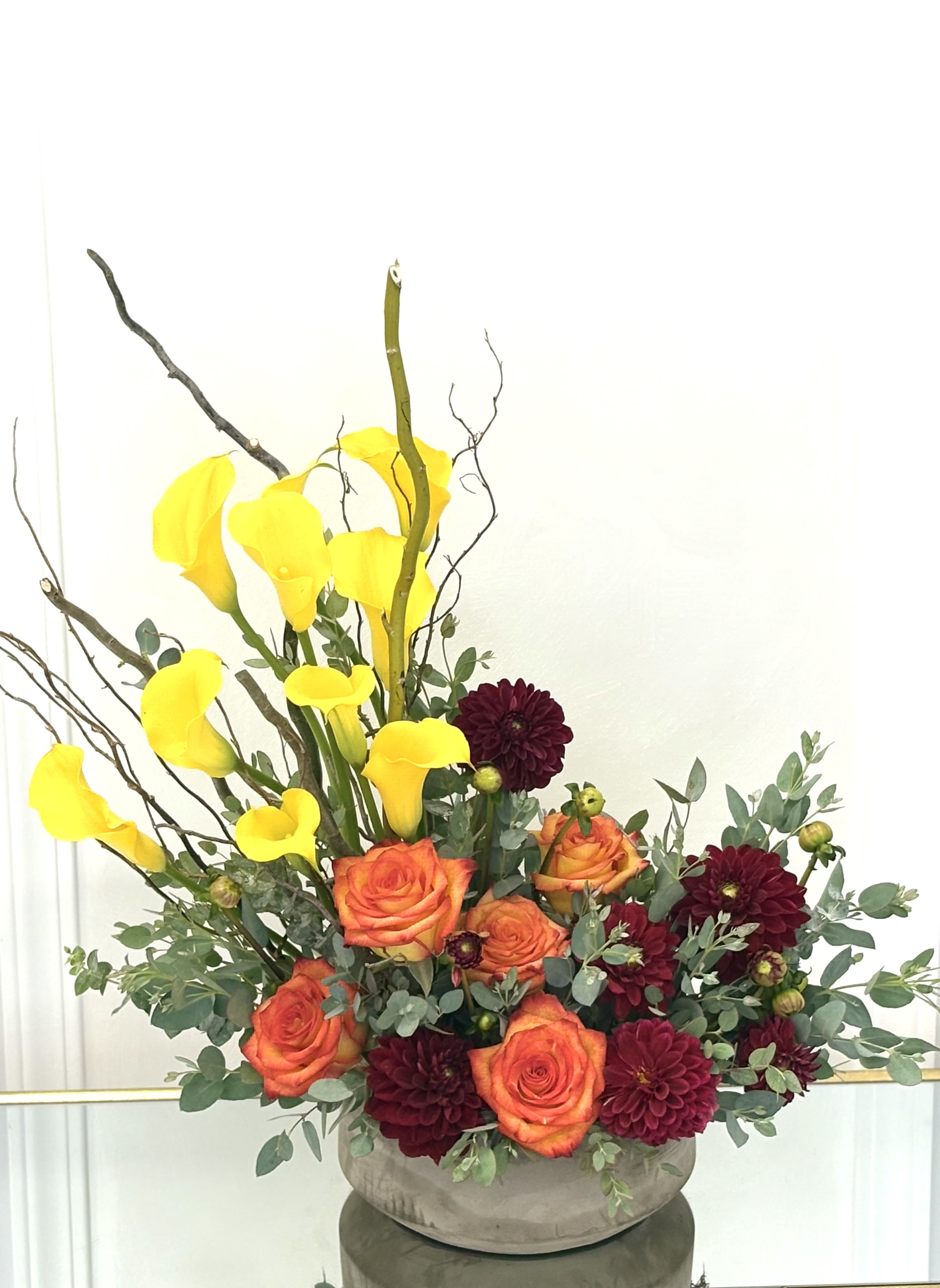 Arrangement of yellow calla lilies, orange roses, and burgundy dahlias in a low vase