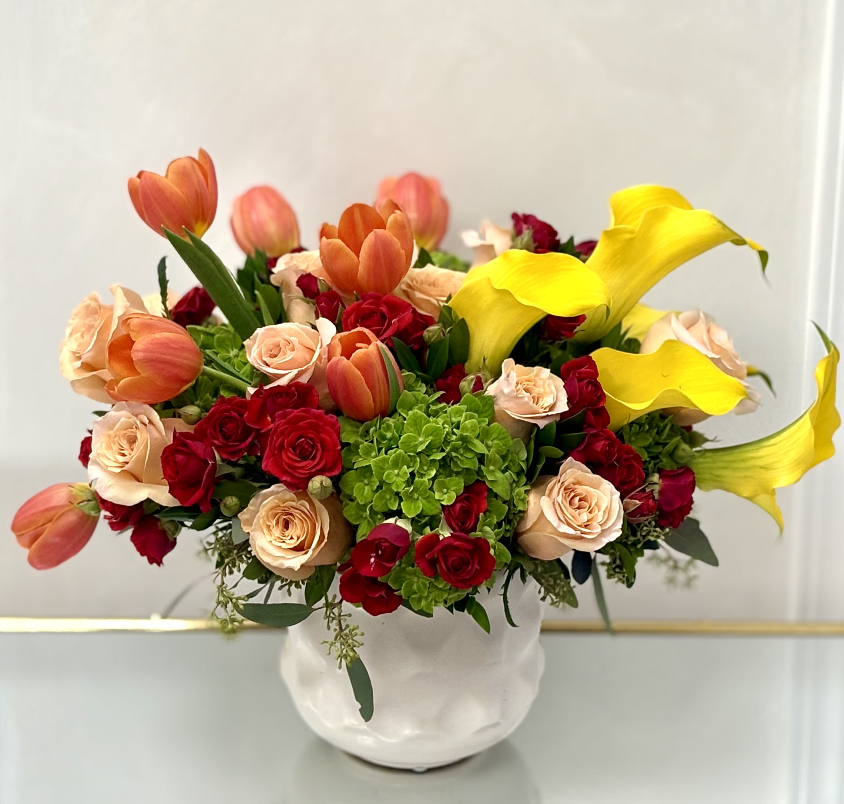 Bouquet of red and peach roses with orange tulips and yellow calla lilies in a white vase