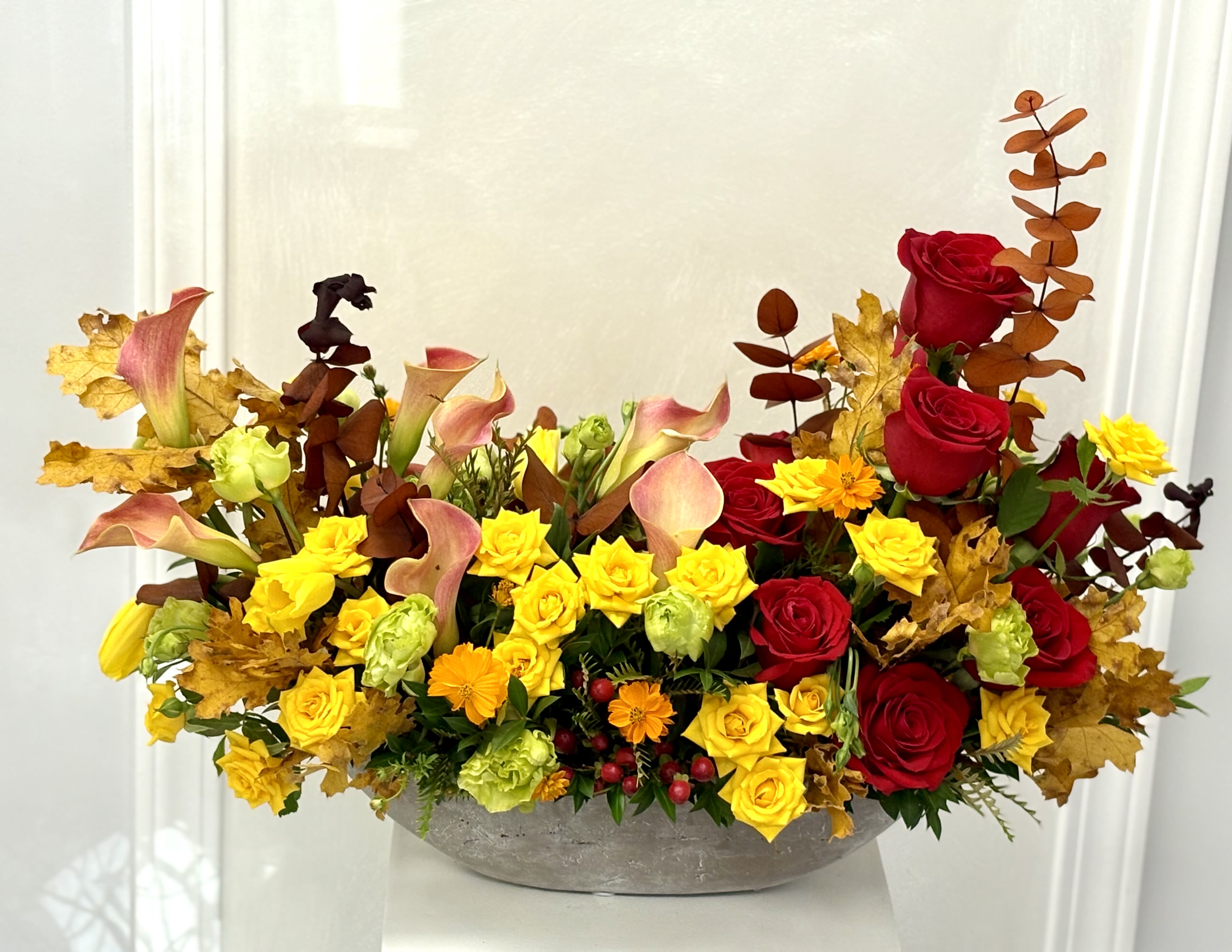 Autumn floral arrangement with red roses, yellow blooms, and calla lilies in a low bowl