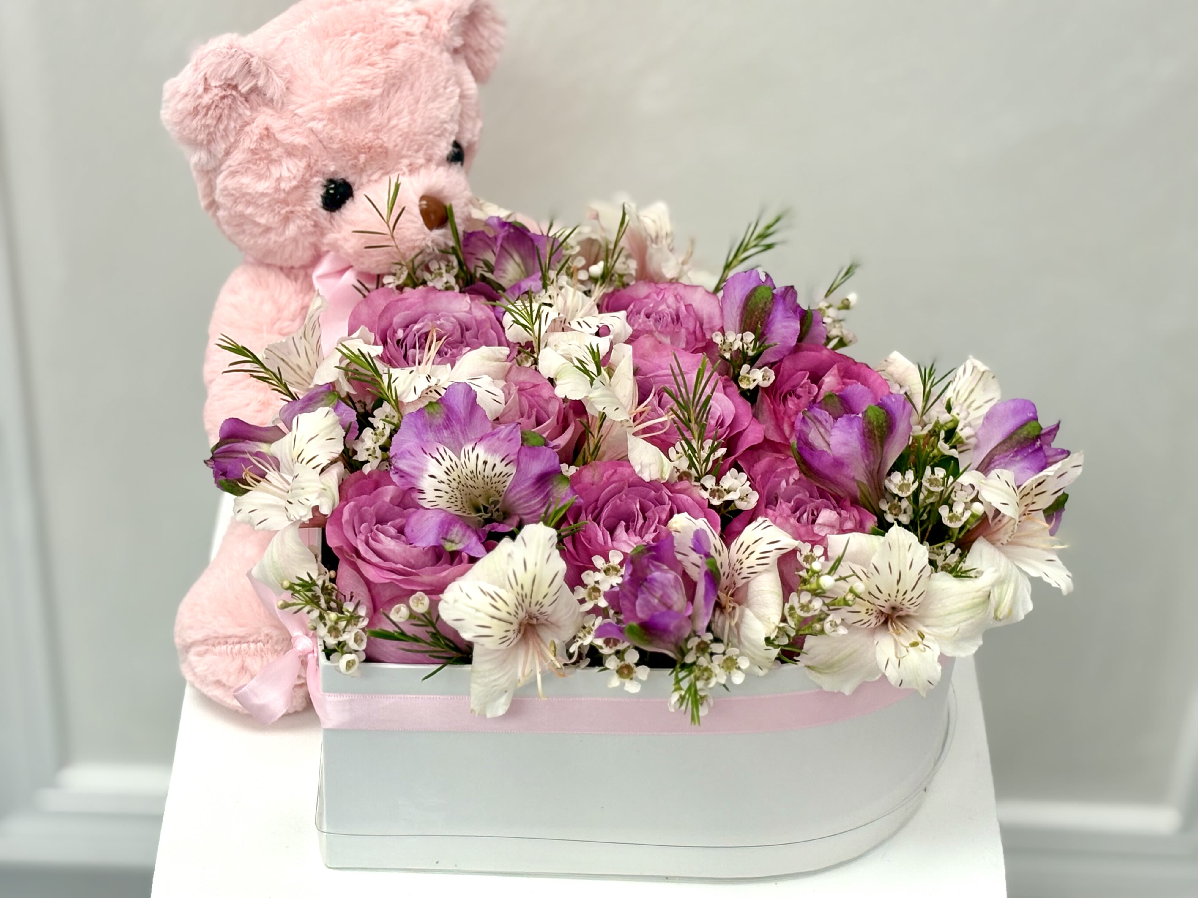 Pink teddy bear hugging a white hat box filled with pink and white flowers