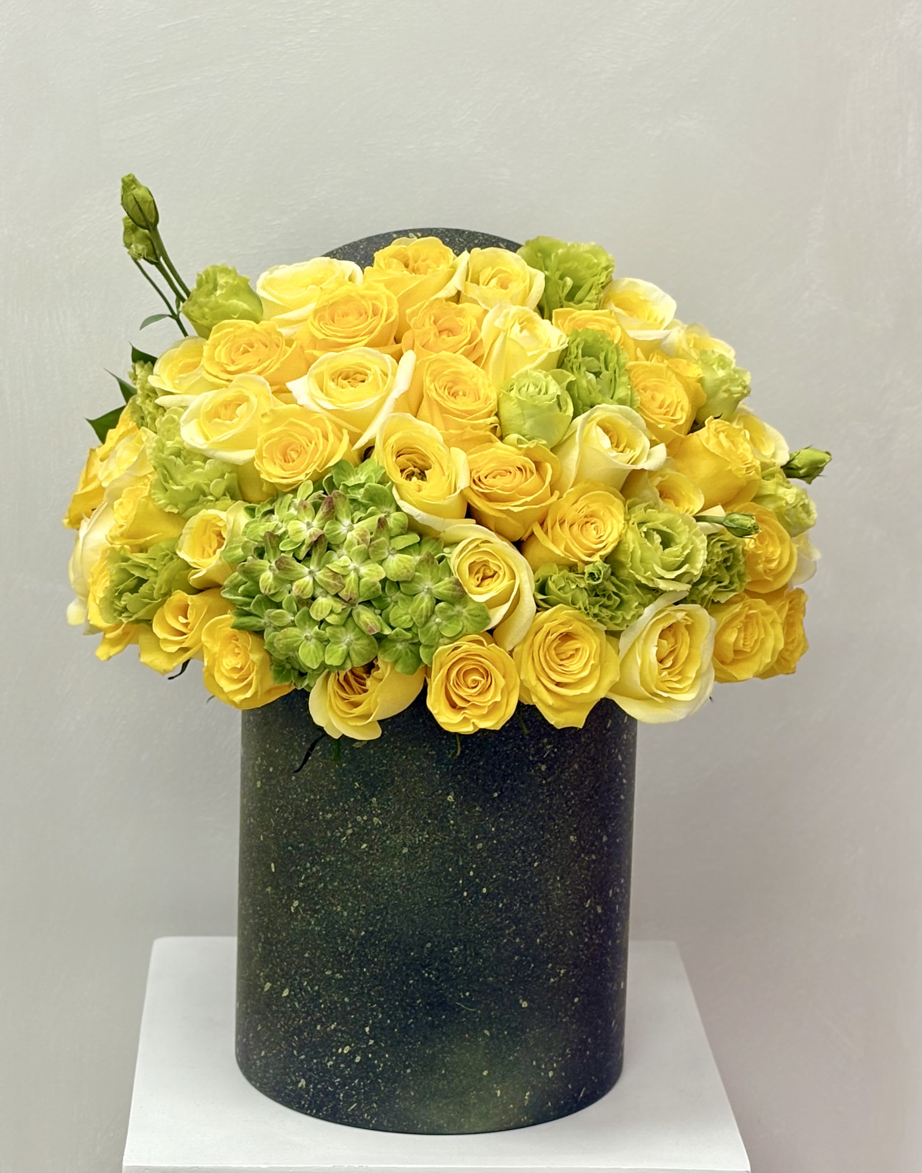 Dense arrangement of yellow and white roses with green blooms in a tall dark green cylinder vase