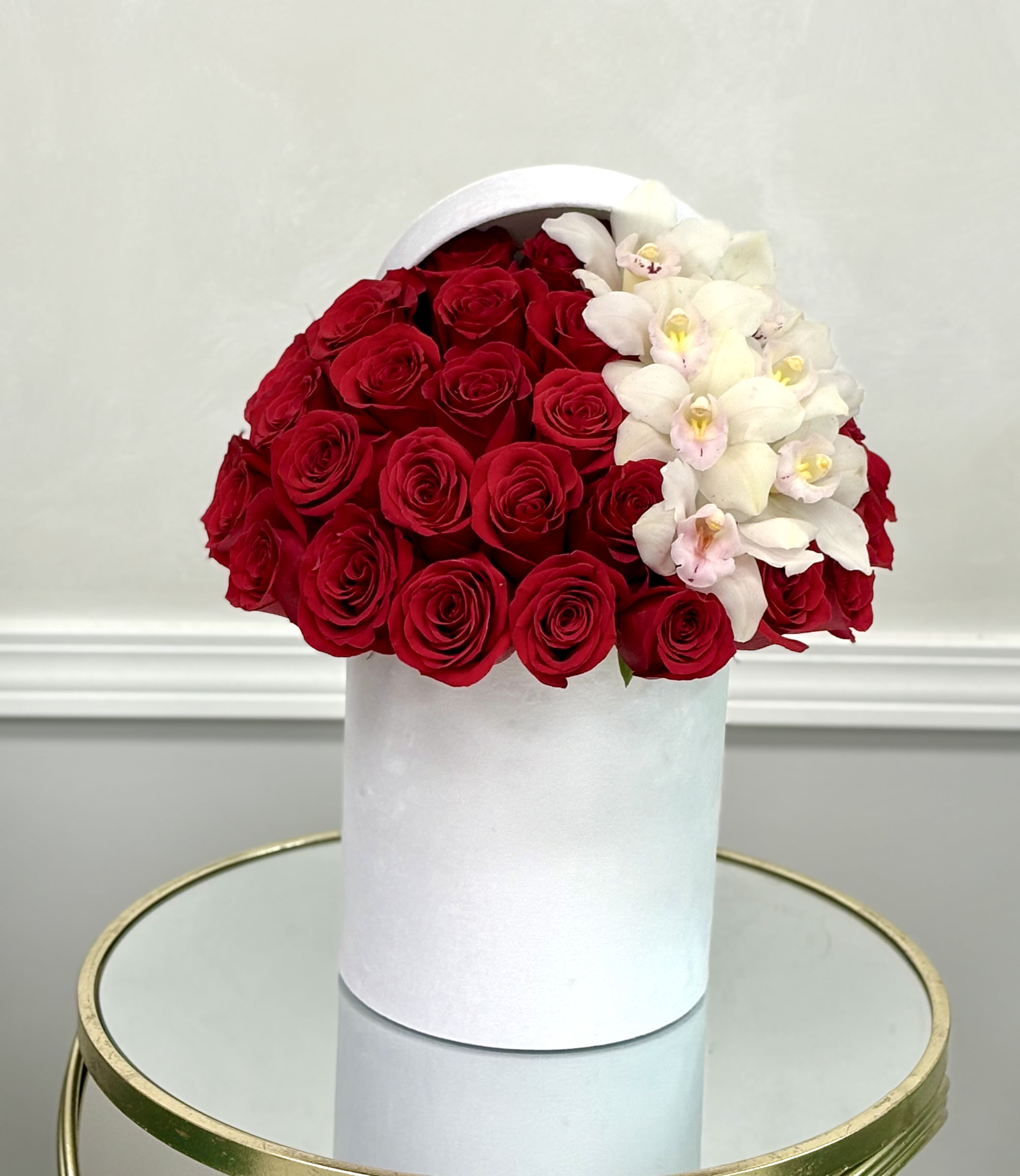 Red roses and white orchids arranged in a white hatbox-style cylinder container on a mirrored table