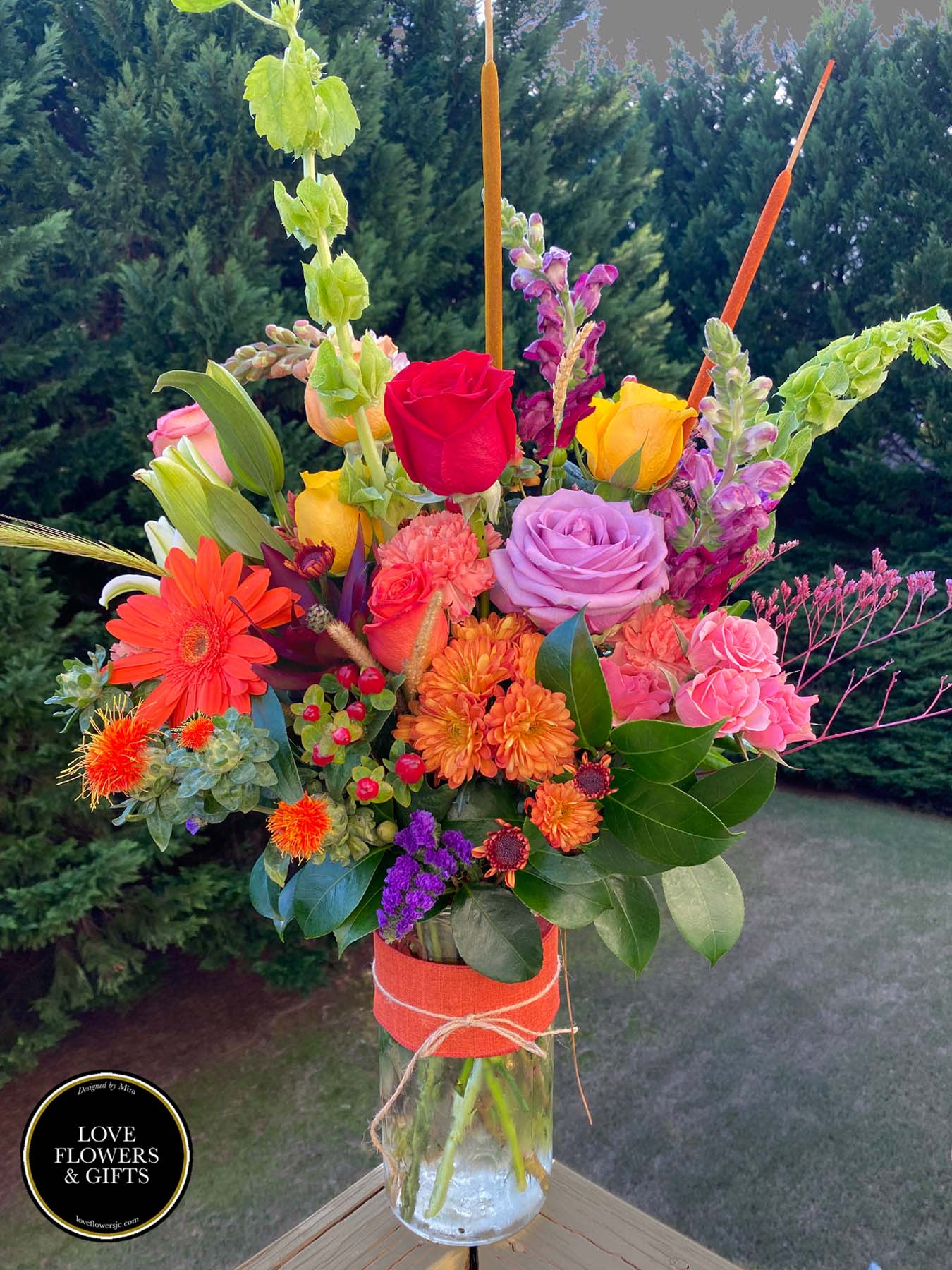 Tall mixed bouquet of bright roses, gerbera daisies, and mums in a glass vase wrapped with orange ribbon.
