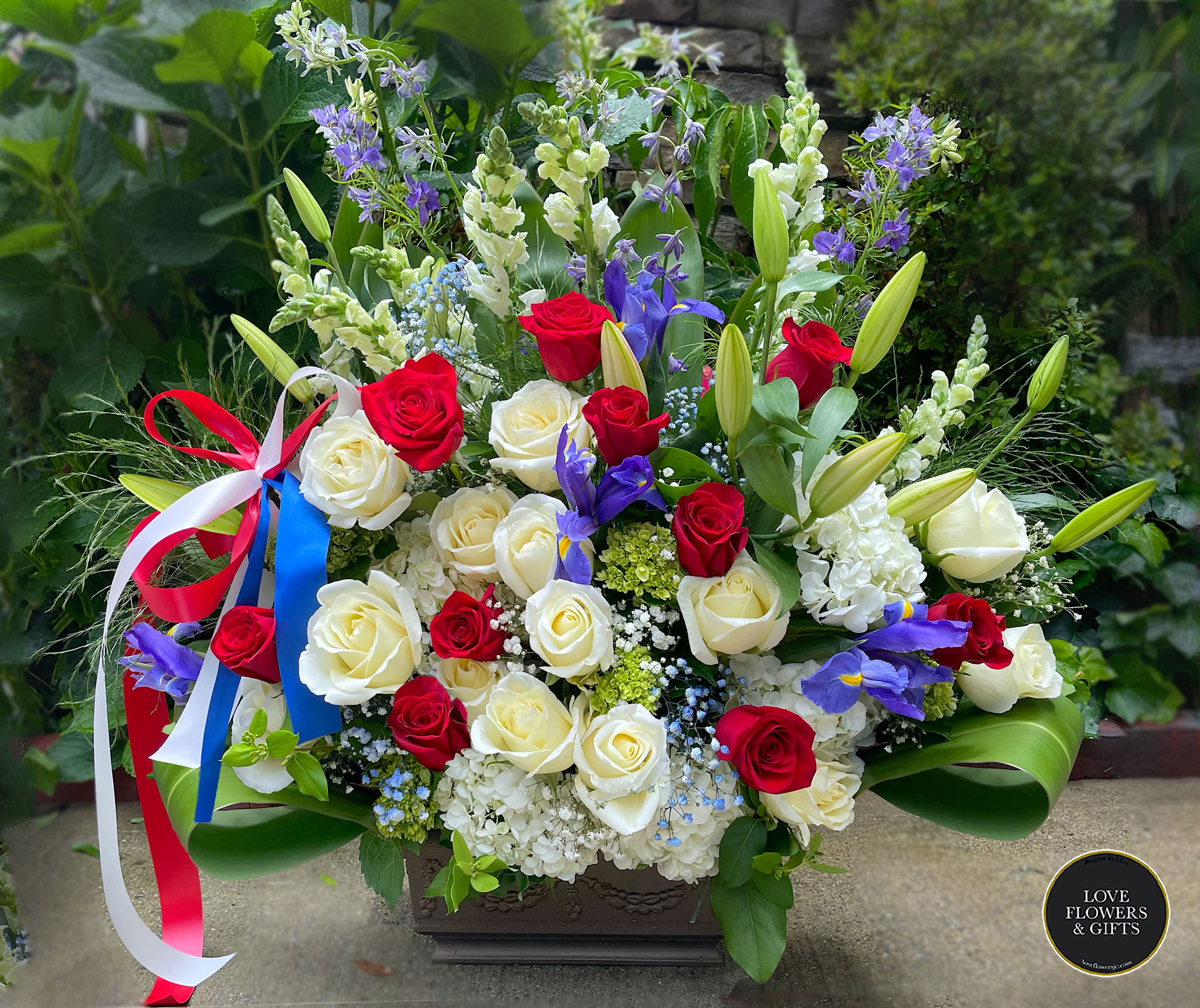 Tall red, white, and blue arrangement with roses, lilies, irises, and a patriotic ribbon in a rectangular container