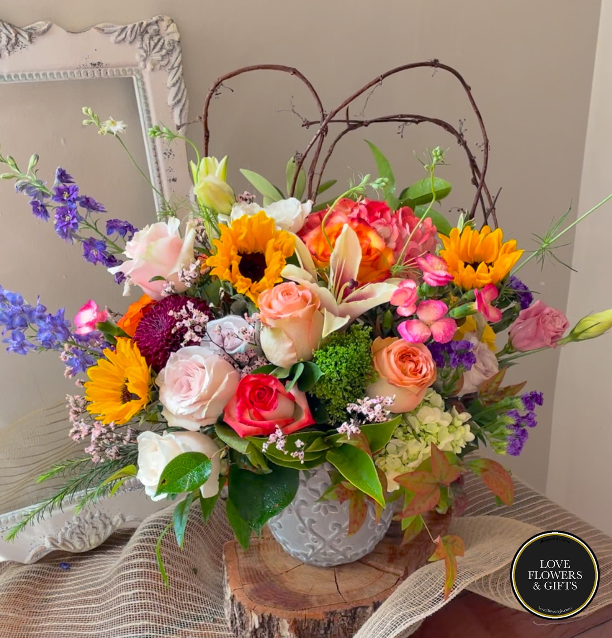 Colorful arrangement of sunflowers, roses, and lilies in a white ceramic pot with twig accents.