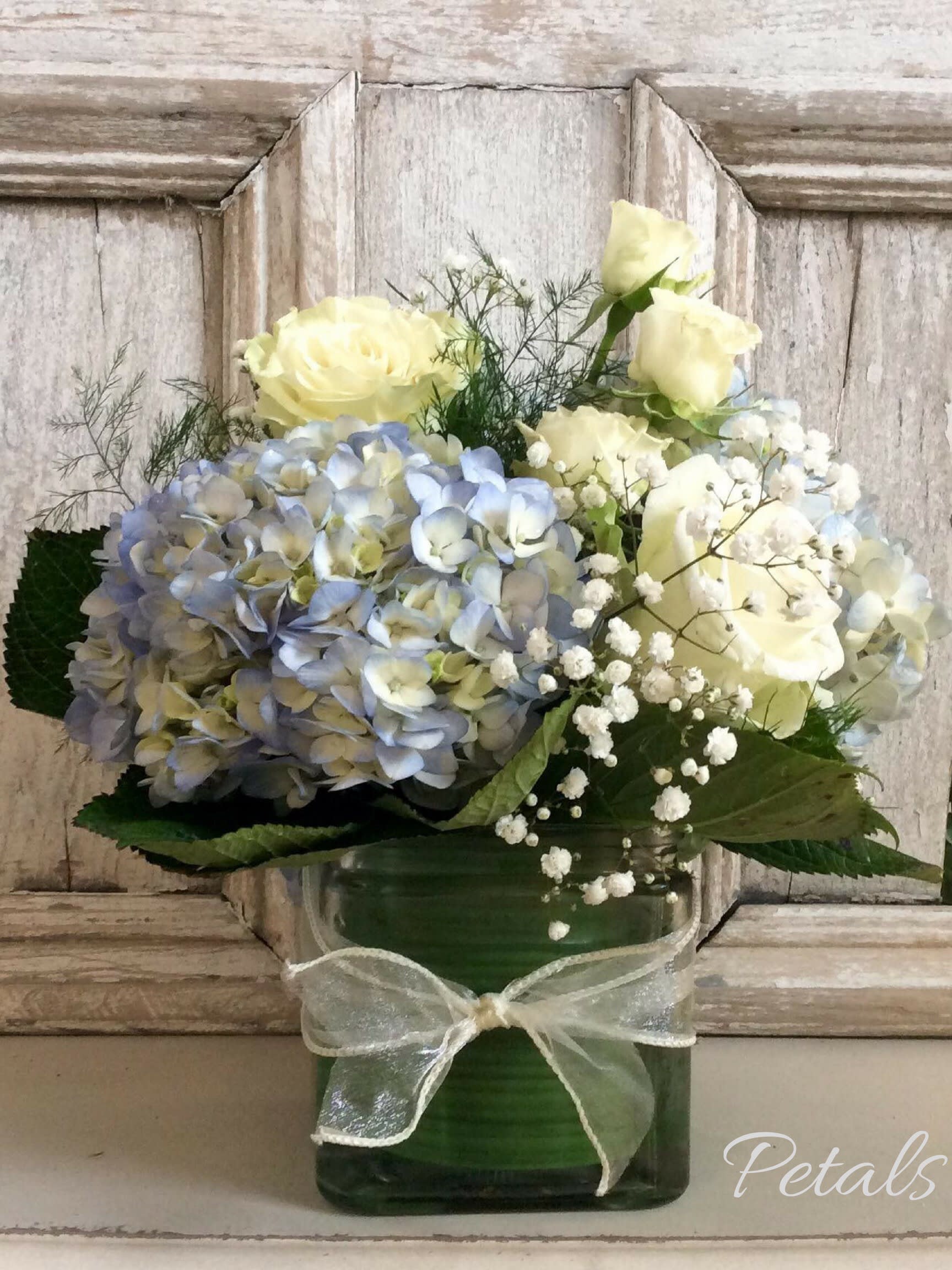 Blue hydrangeas and pale yellow roses in a green glass vase with a ribbon