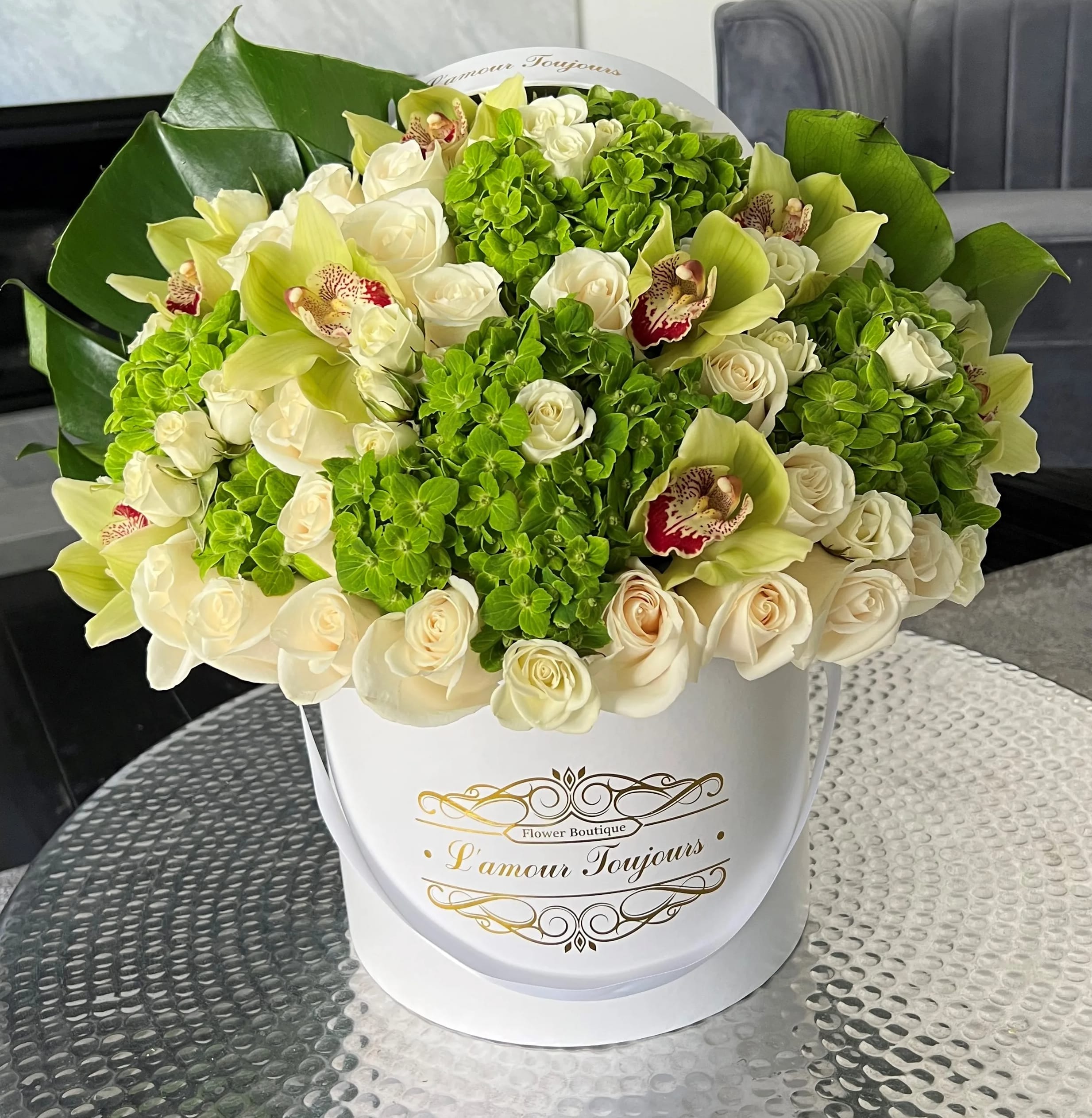 Round box arrangement of cream roses, green hydrangeas, and green orchids on a glass table