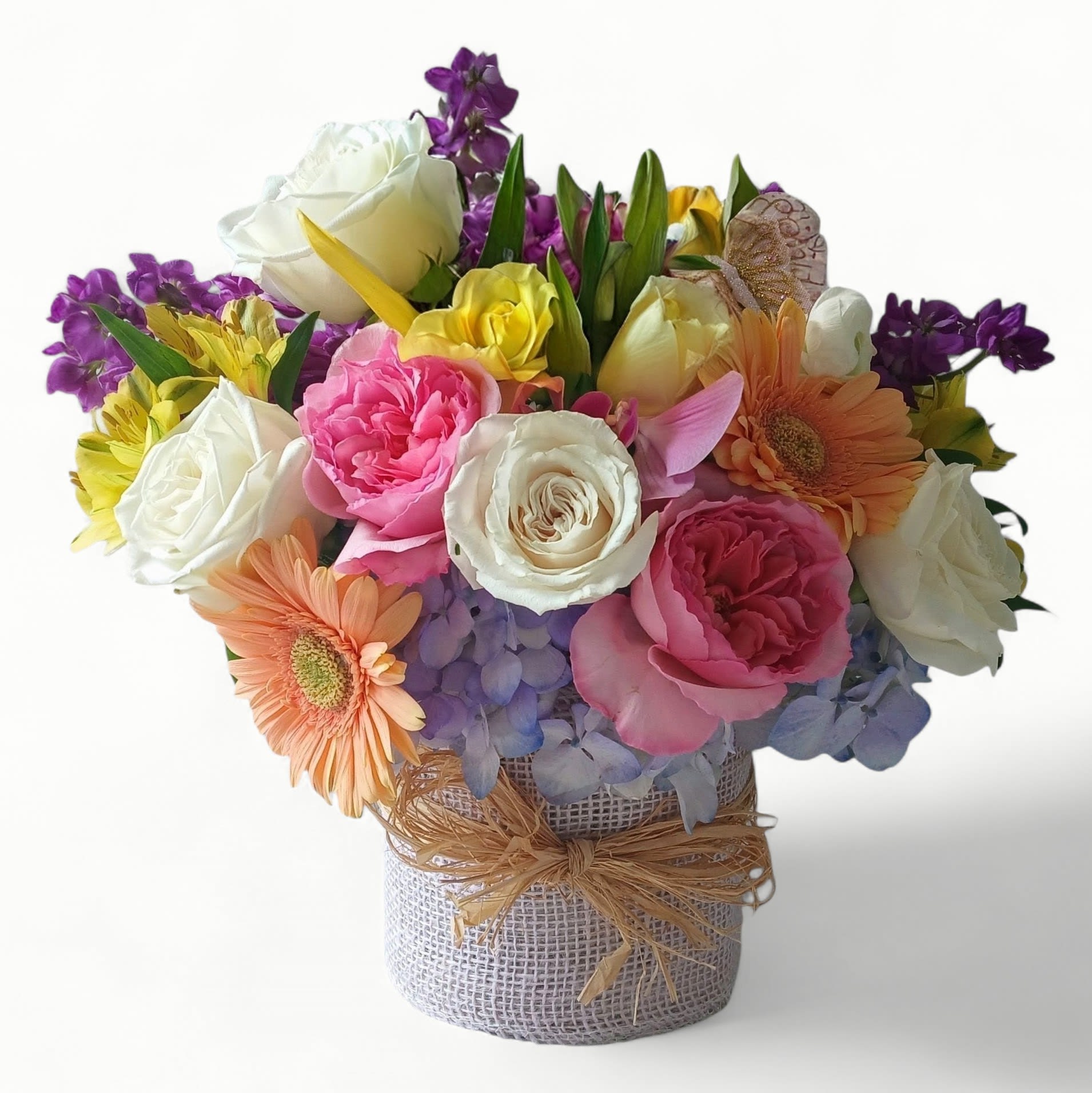 Happy Birthday Blossom Bouquet Designer's Choice - Bright pastel blooms packed into a burlap-wrapped vessel create an instant "happy birthday" surprise that feels both cheerful and gently romantic. This flower arrangement for Birthday celebrations features roses and hydrangeas up front, with hints of stock and gerbera daisies in pink, purple, white, yellow, peach, and soft blue-an uplifting mix that makes the recipient feel truly celebrated. The dense, rounded shape reads like a joyful bouquet in a keepsake container, ideal for summer parties, housewarmings, or retirement gatherings.  Carefully arranged in a rustic burlap-wrapped vase tied with a raffia bow, the Happy Birthday Blossom Bouquet Designer's Choice is made to look hand-tied while sitting on a table or mantel. Expect closely packed white and pink roses, soft blue hydrangeas, peach and yellow gerbera-style accents, and complementary purple stock for texture and scent. The design arrives ready to display; change water every other day, recut stems if you transfer to another vase, and keep away from direct heat to extend vase life. Local delivery available to Haddonfield, NJ, for same-day gifting that brightens a birthday, summer celebration, housewarming, or milestone retirement.