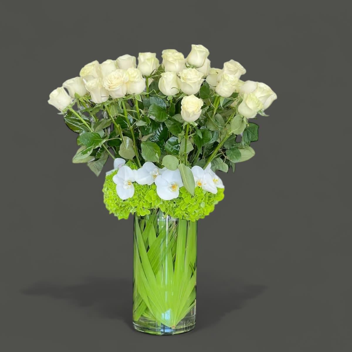 White roses and orchids in a tall clear vase with bright green flowers